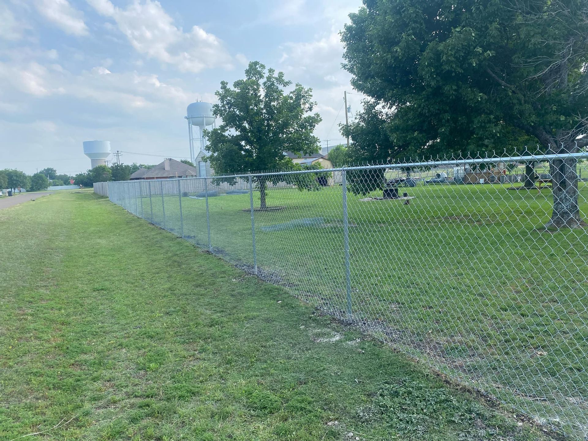 Chain-link fence along a grassy park field under a cloudy sky, with trees and distant houses.