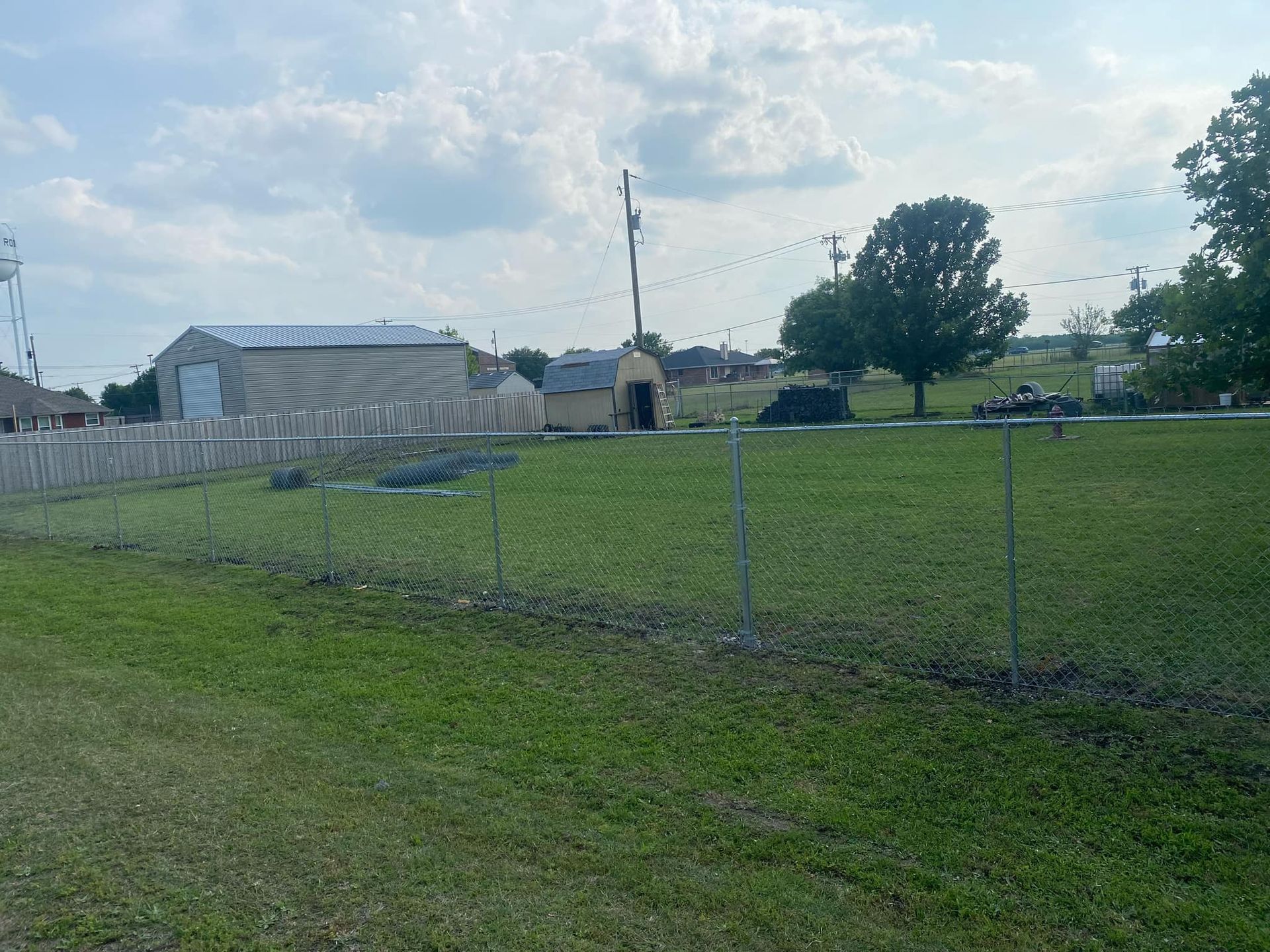 Grassy field with a chain-link fence, greenhouse, and trees under a cloudy sky.
