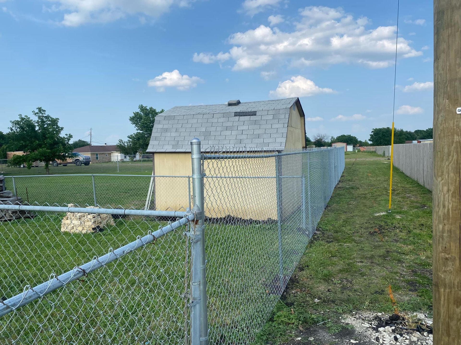 Chain-link fence and small shed in a grassy yard under a blue sky with clouds