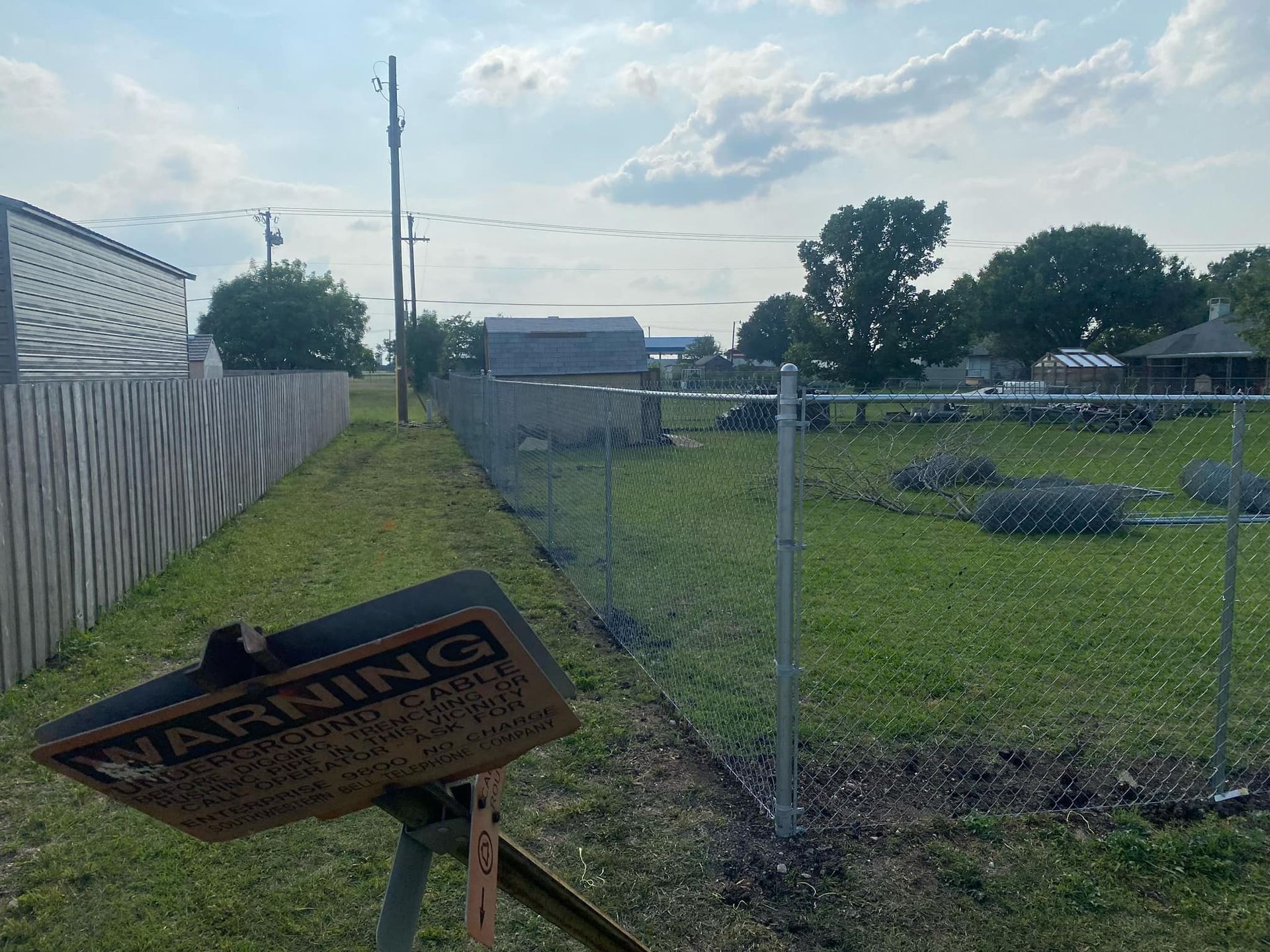 Damaged warning sign beside a chain-link fence and grassy field under a cloudy sky