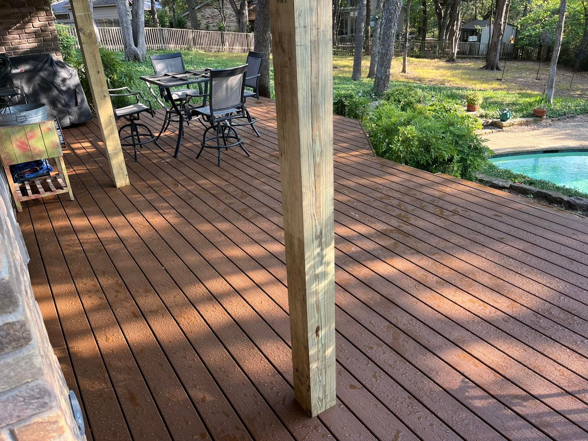 Wooden deck with patio chairs, a central post, and a pool beside landscaped greenery