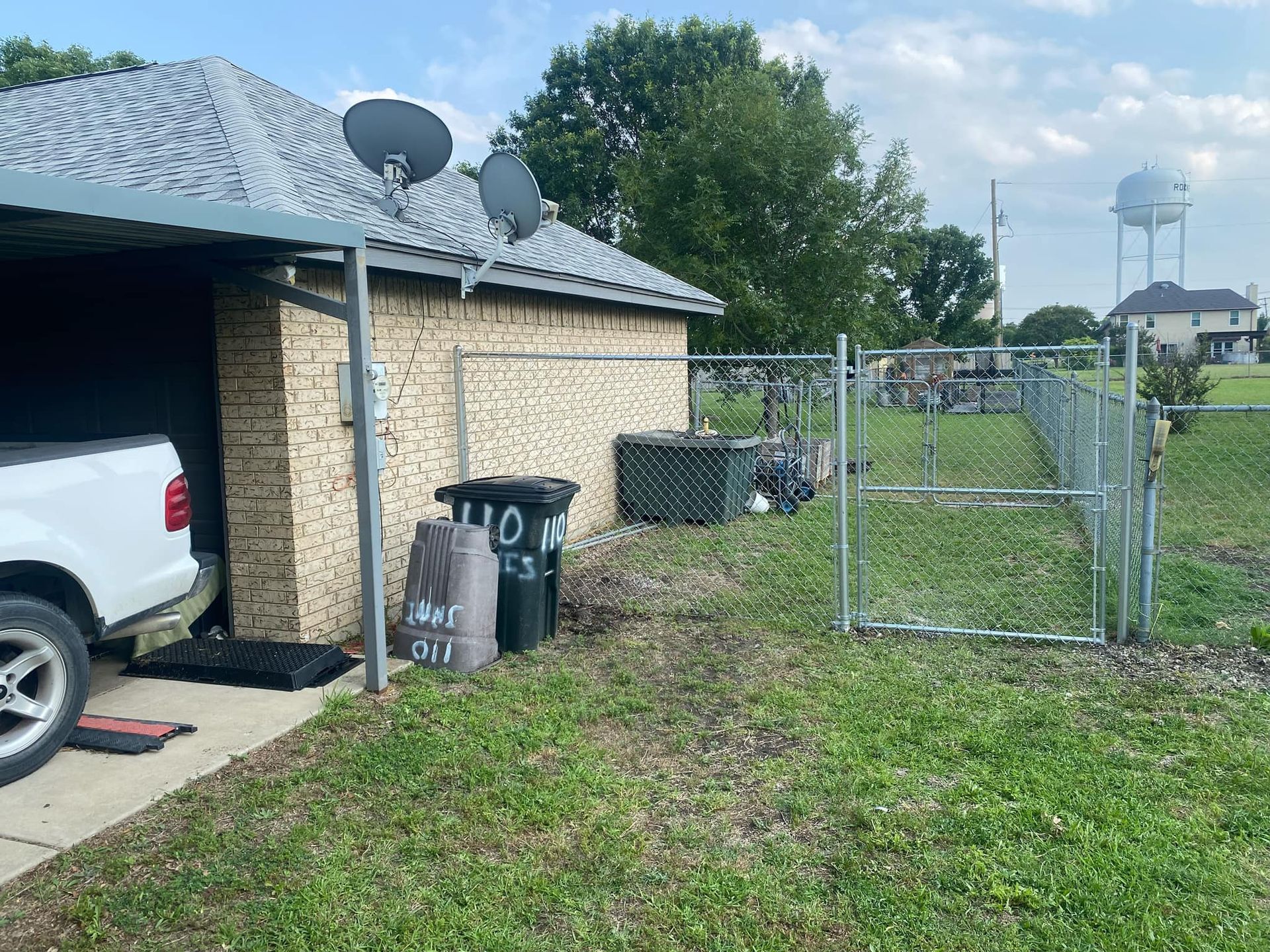 Backyard beside brick house and garage, with white SUV, trash bins, chain-link fence, and water tower in distance