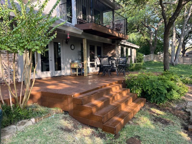 Wooden deck and stairs leading to a shaded backyard patio beside a house, with trees and greenery.