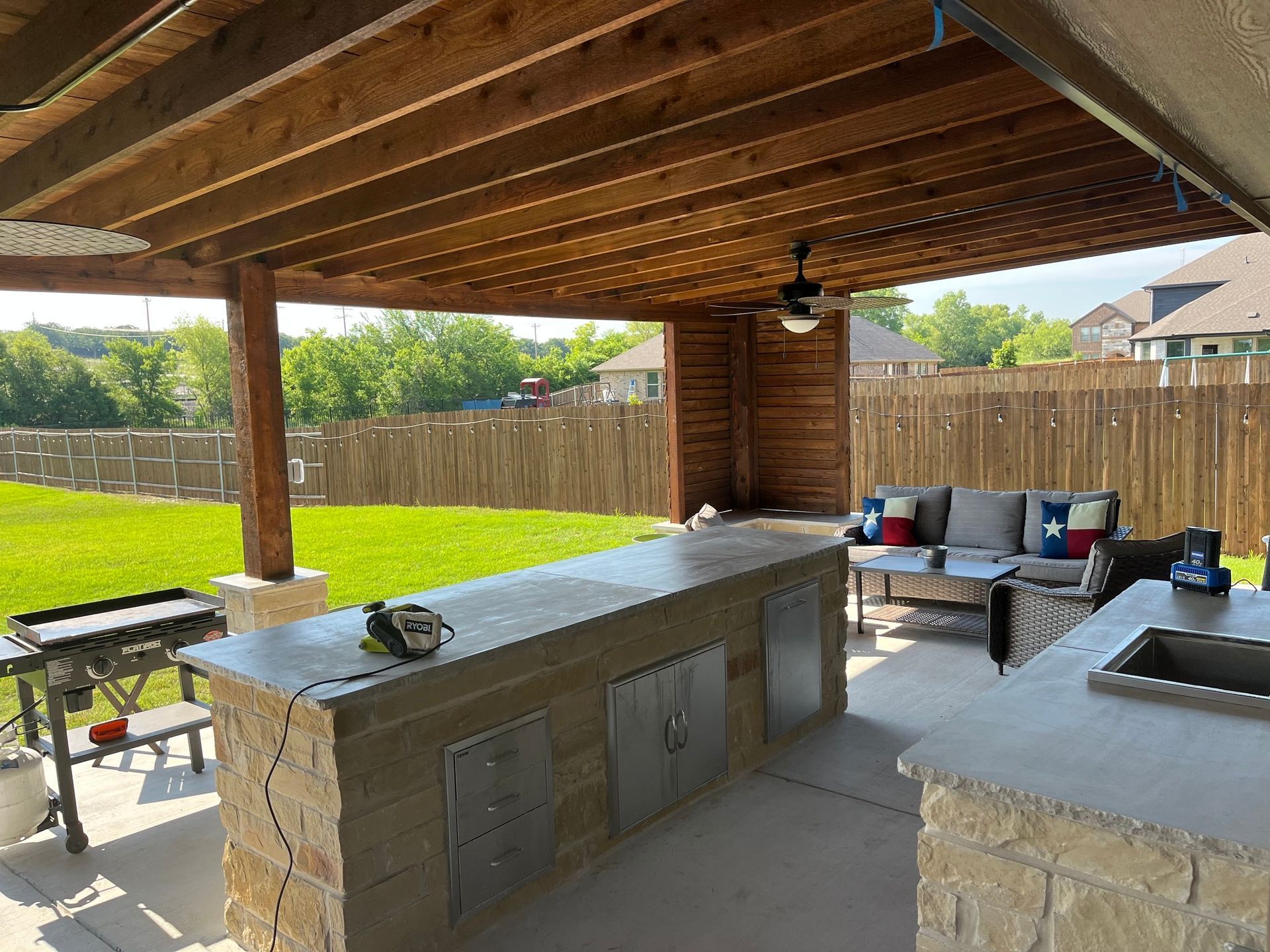 Covered backyard patio with outdoor kitchen, seating, and a grassy yard bordered by a wooden fence.