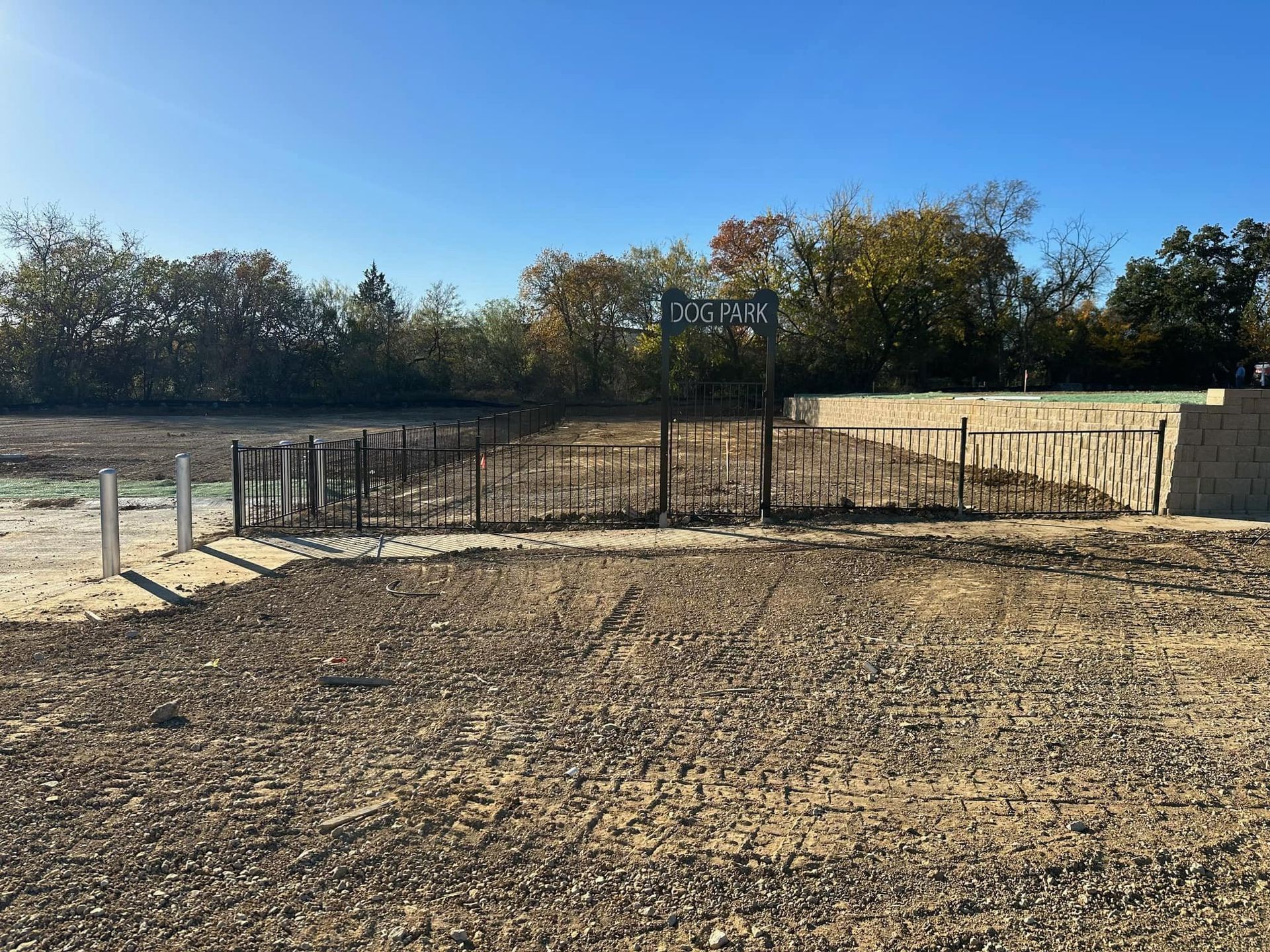 Empty dirt lot with a fenced area, stone wall, and trees under a clear blue sky