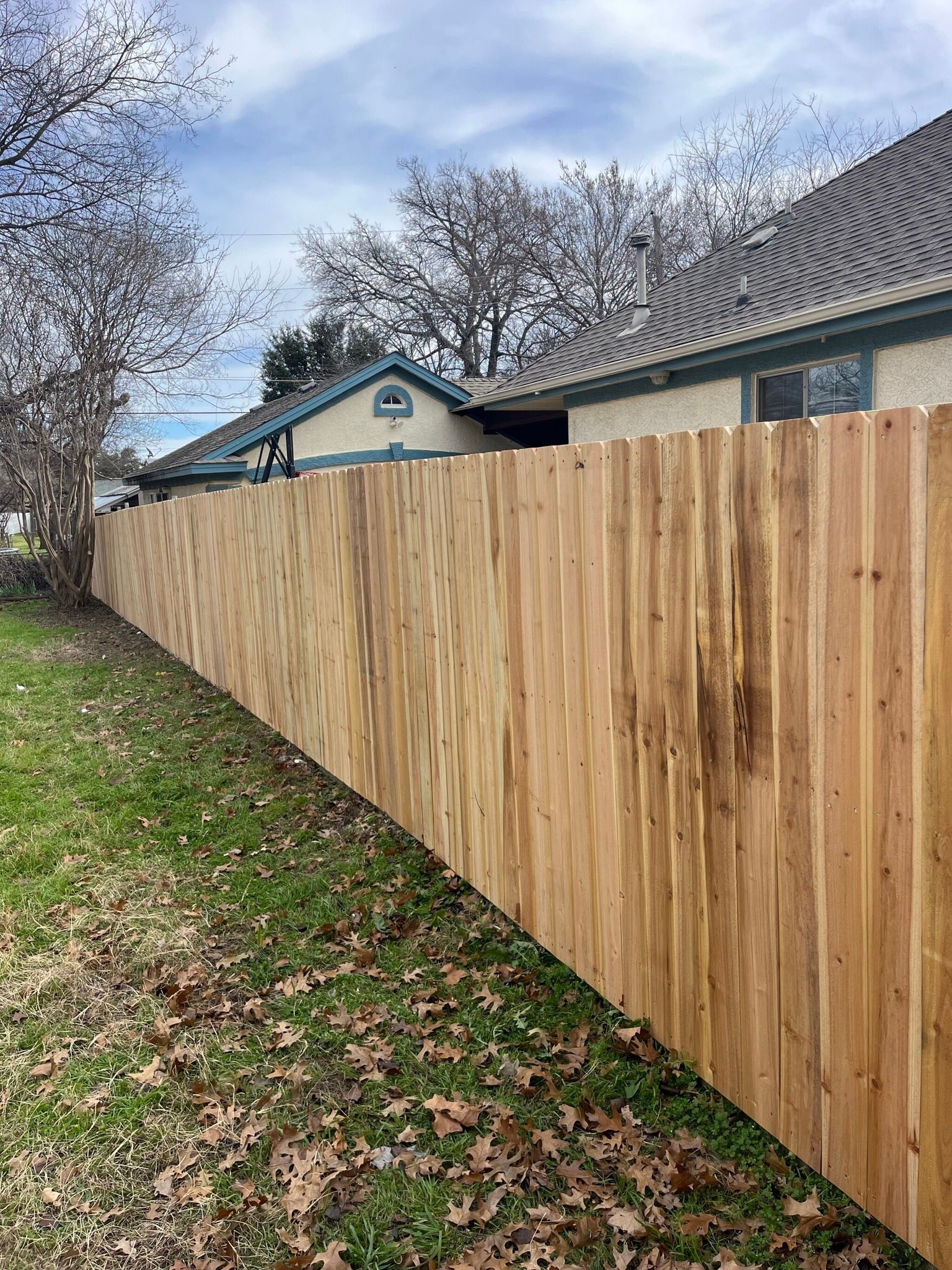 Wooden fence beside a house with a gray roof, along a leaf-covered yard and cloudy sky