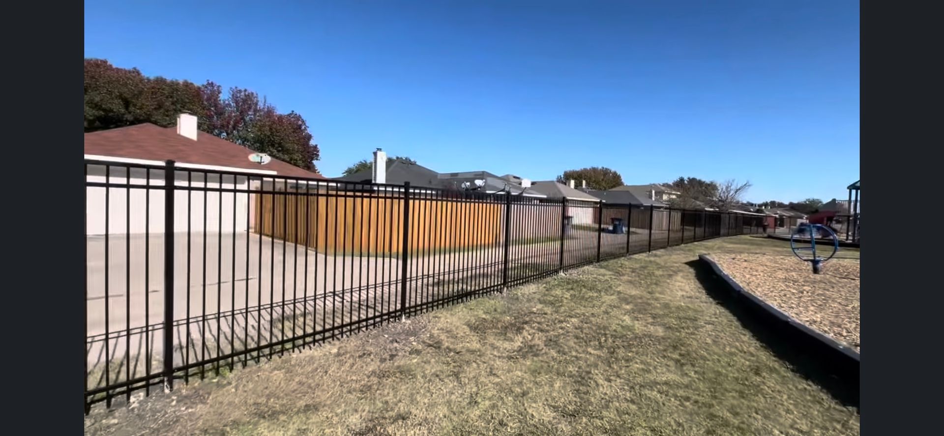 Residential backyard with a long black fence beside a tan wall under a clear blue sky