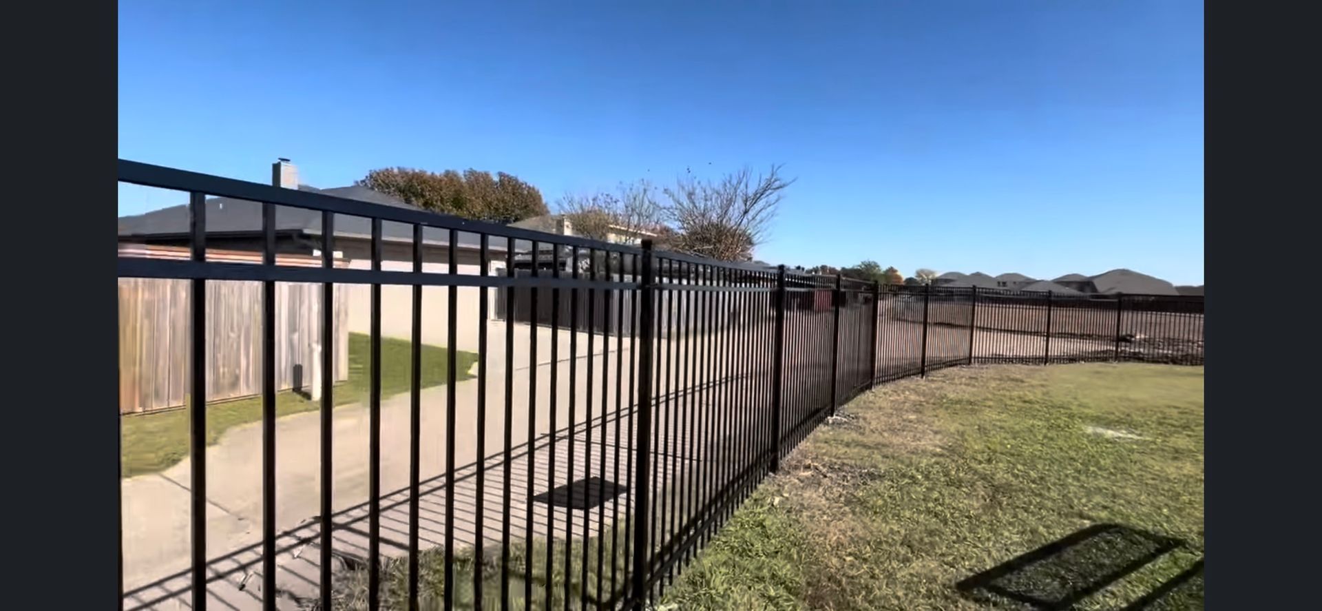Black metal fence beside a tan wooden fence along a grassy yard under a clear blue sky