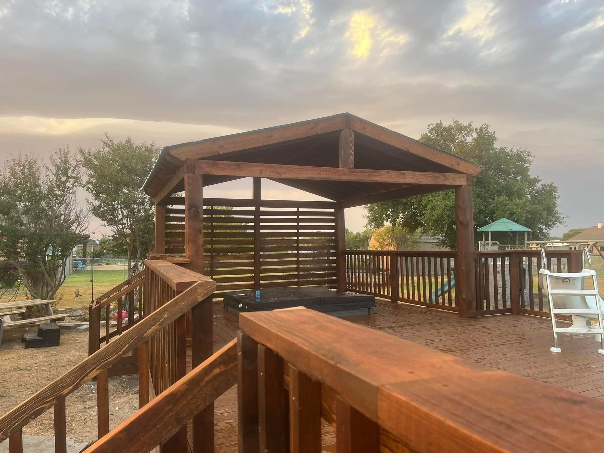 Wooden pavilion and ramped deck in a park at sunset, with trees and playground equipment nearby