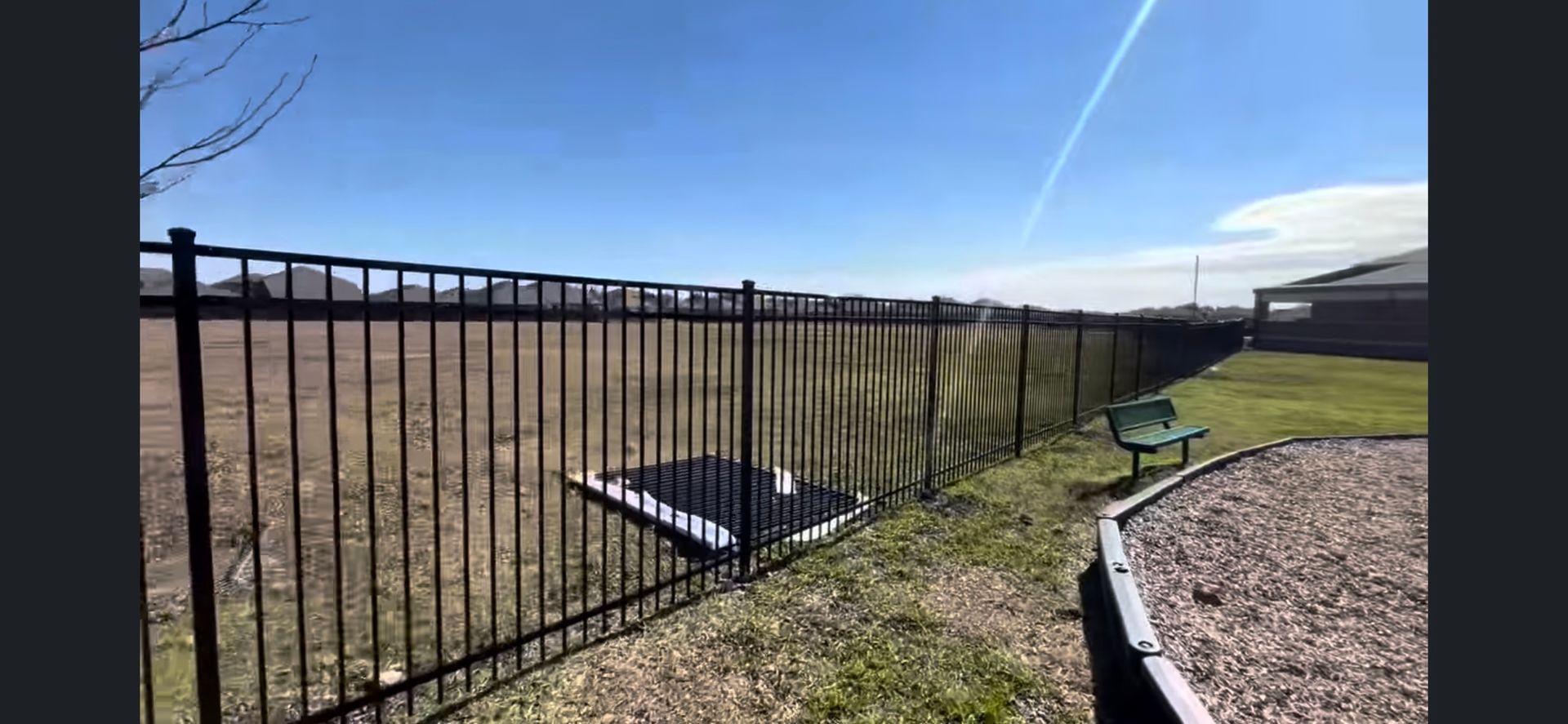 Black metal fence around a grassy yard with a solar panel on the ground under a bright blue sky.