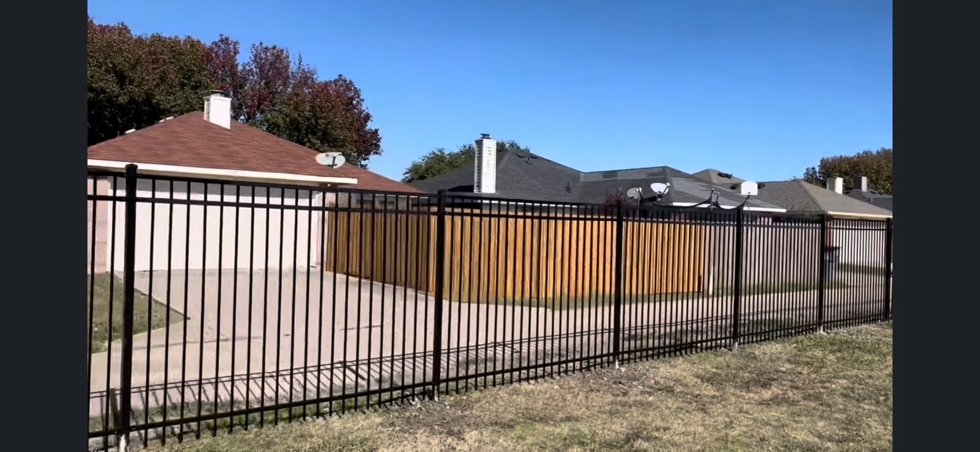 Residential backyard with a black metal fence, wooden privacy fence, and single-story houses under a blue sky