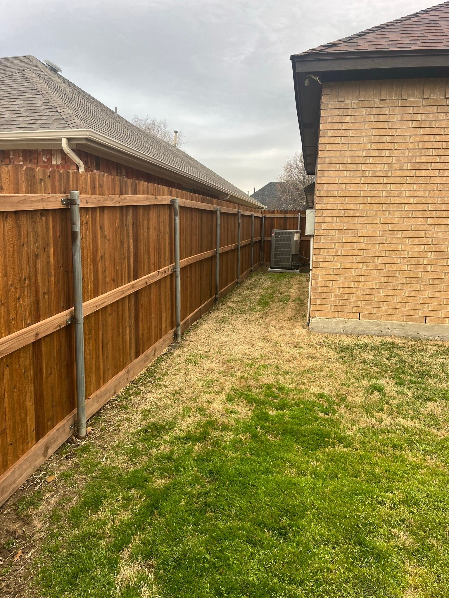 Narrow grassy side yard between a wooden fence and a brick house under an overcast sky