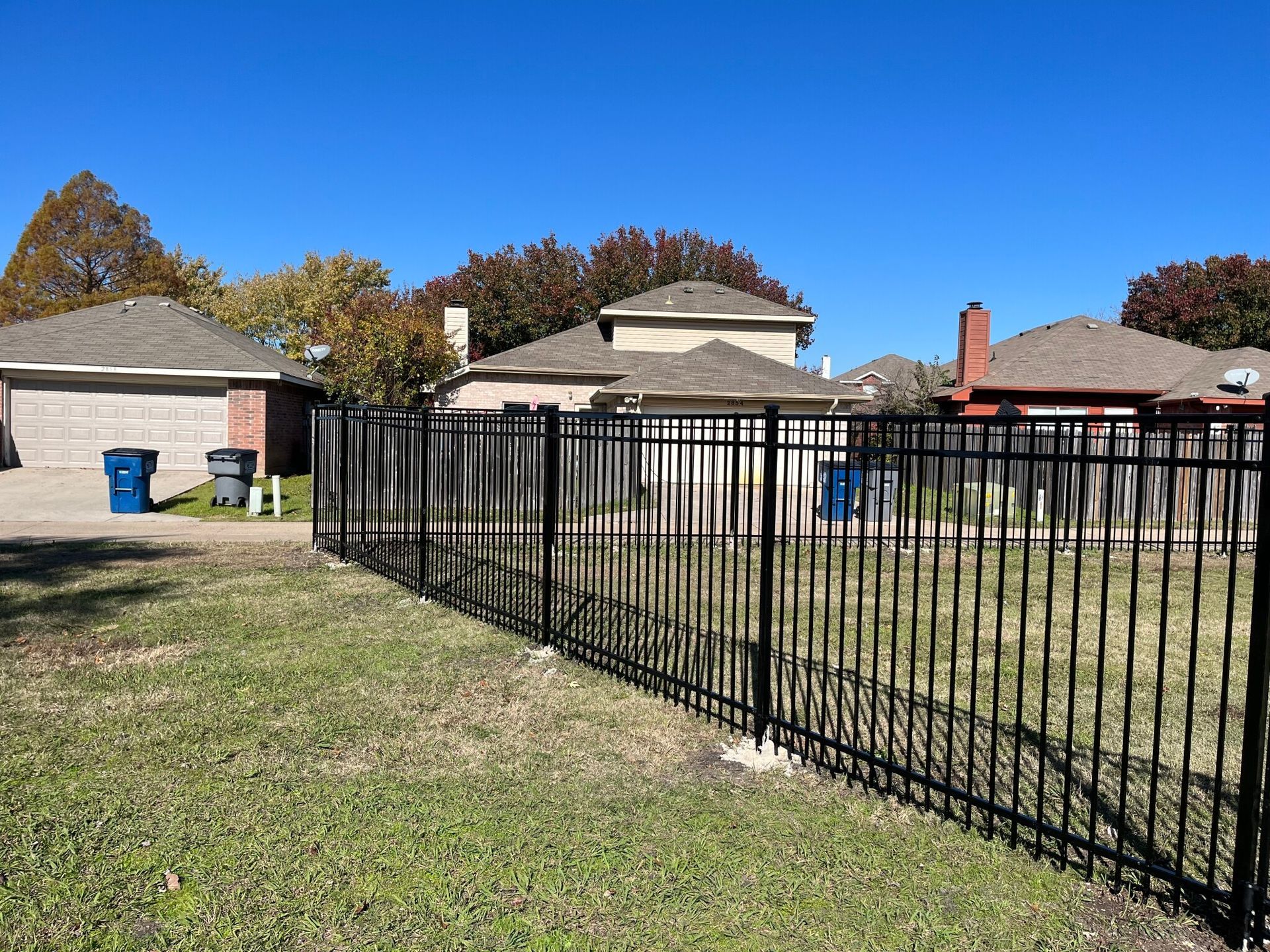 Black metal fence along a grassy yard beside single-story houses under a clear blue sky.