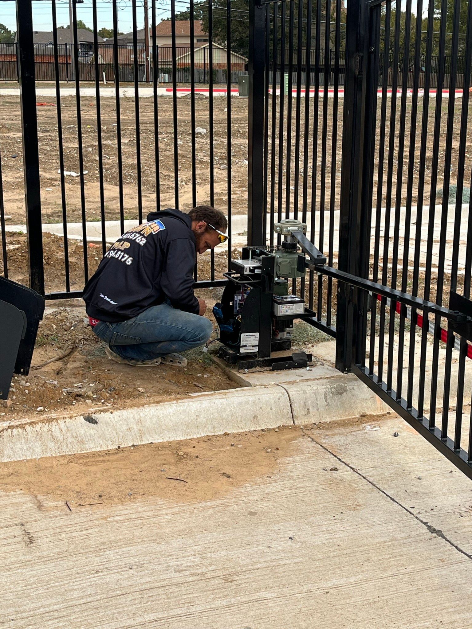 Person kneeling by a fence, inspecting a utility meter or valve box outdoors.