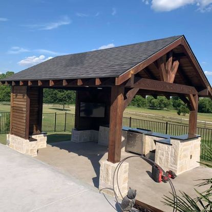 Wooden pavilion with stone bases and a shingled roof on a concrete patio beside a grassy field