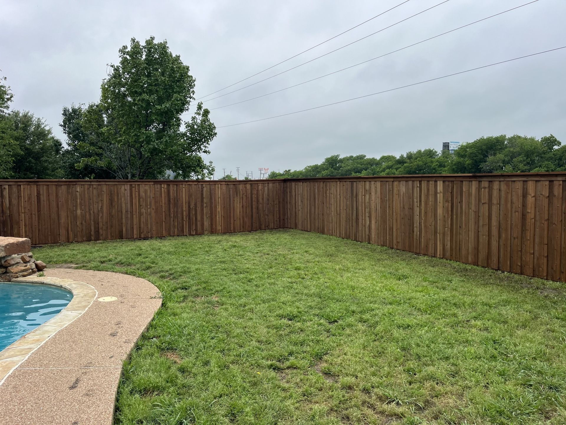 Backyard with grassy lawn, wooden privacy fence, and pool edge under cloudy sky