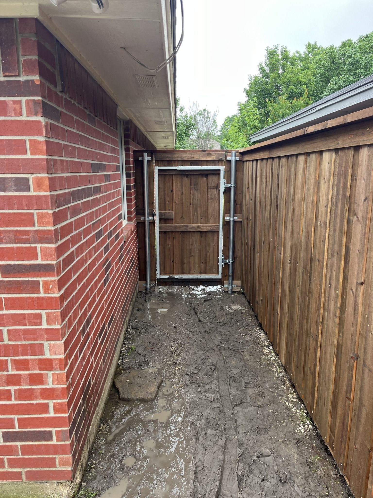 Narrow muddy side yard between a brick house and wooden fence, ending at a wooden gate.