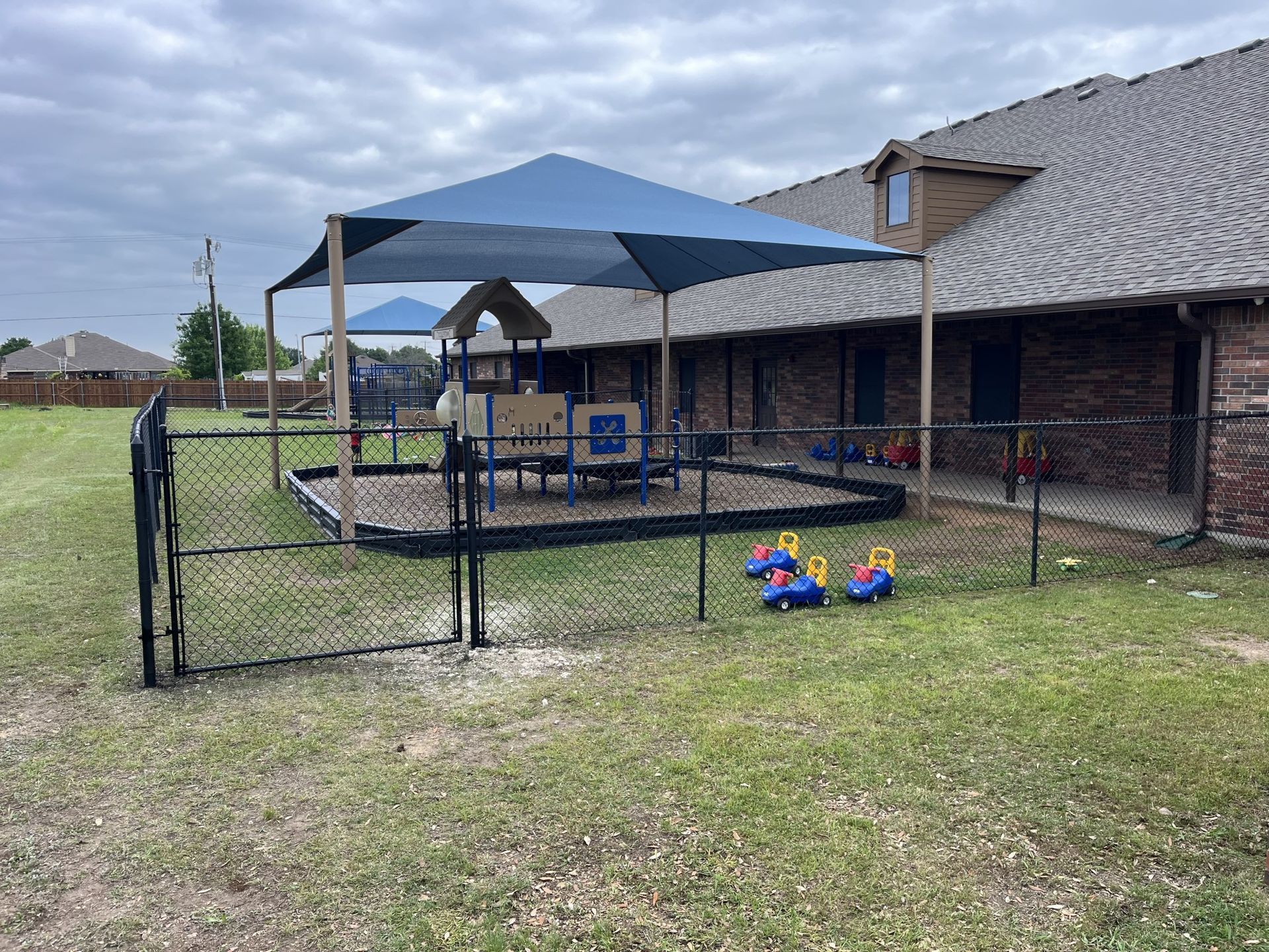 Backyard patio with blue canopy, fenced grassy yard, and scattered toys beside a brick house