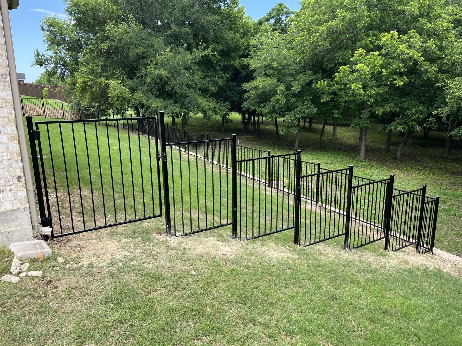 Black metal fence along a grassy slope beside trees and a stone wall in a sunny yard