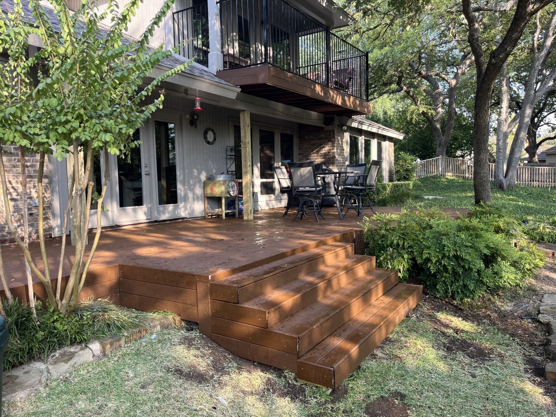 Two-story house with wooden deck steps and patio seating in a shaded yard