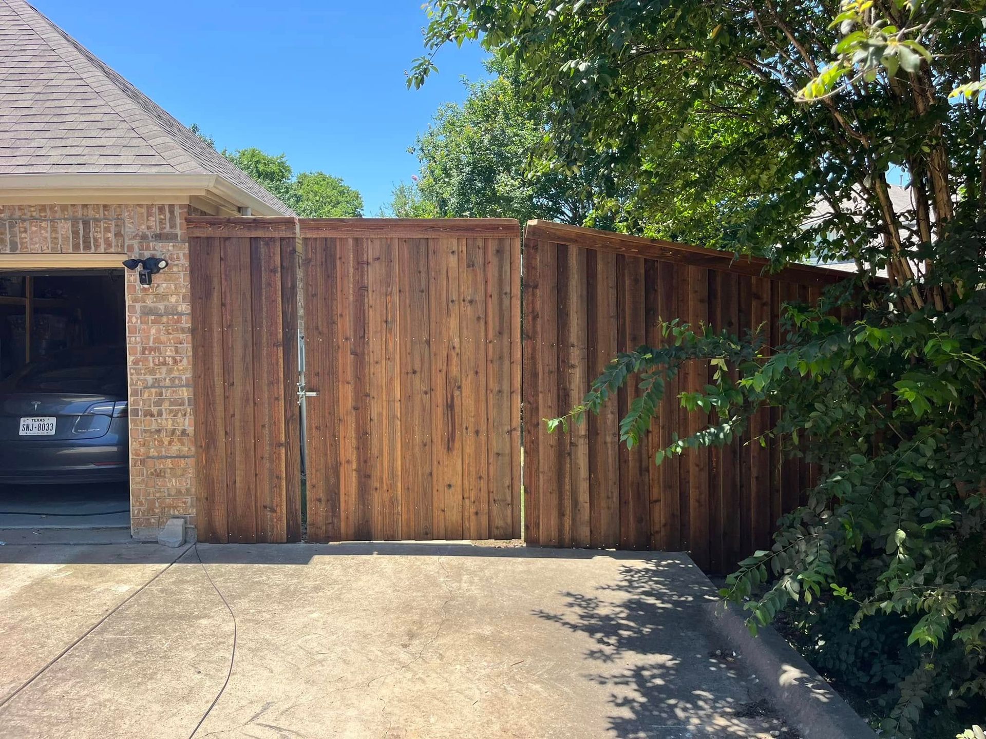 Wooden driveway gate beside a garage, with trees and a clear blue sky overhead.