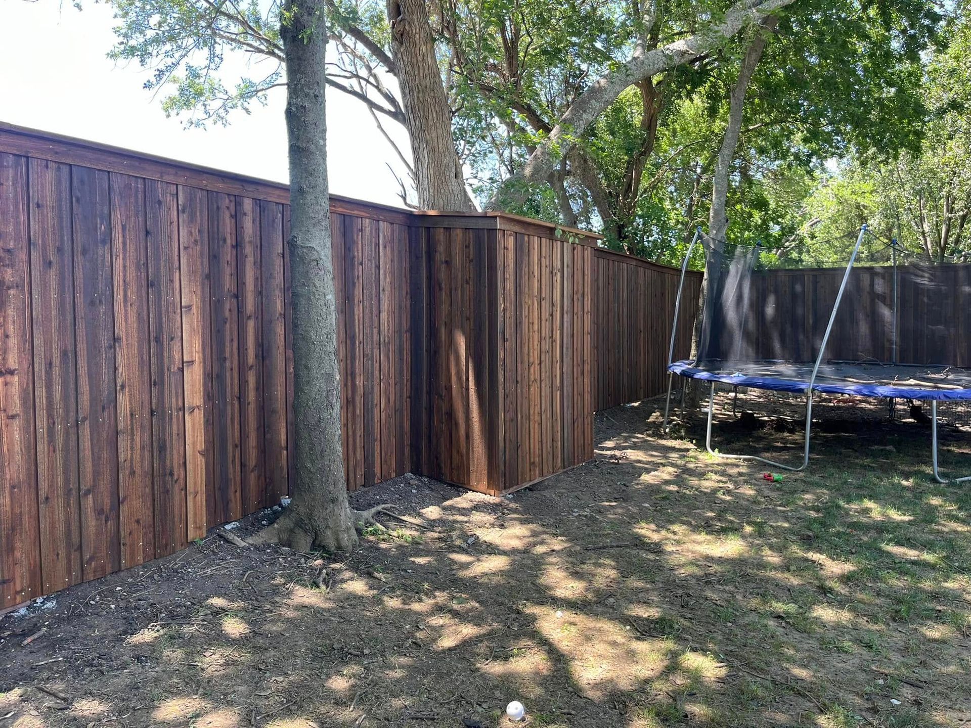 Backyard with wooden fence, trees, and a trampoline on a grassy lawn.