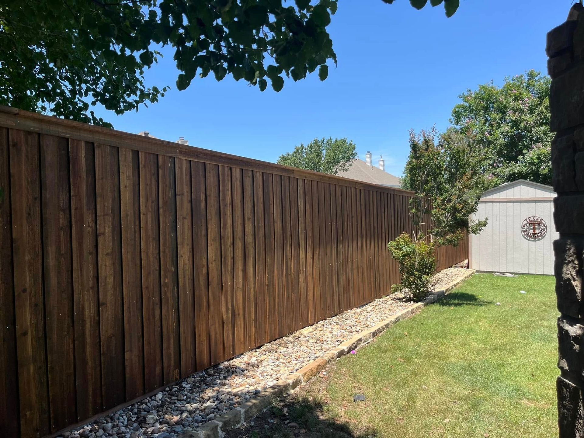 Backyard with a tall wooden fence, green lawn, stone border, and a small white shed under blue sky