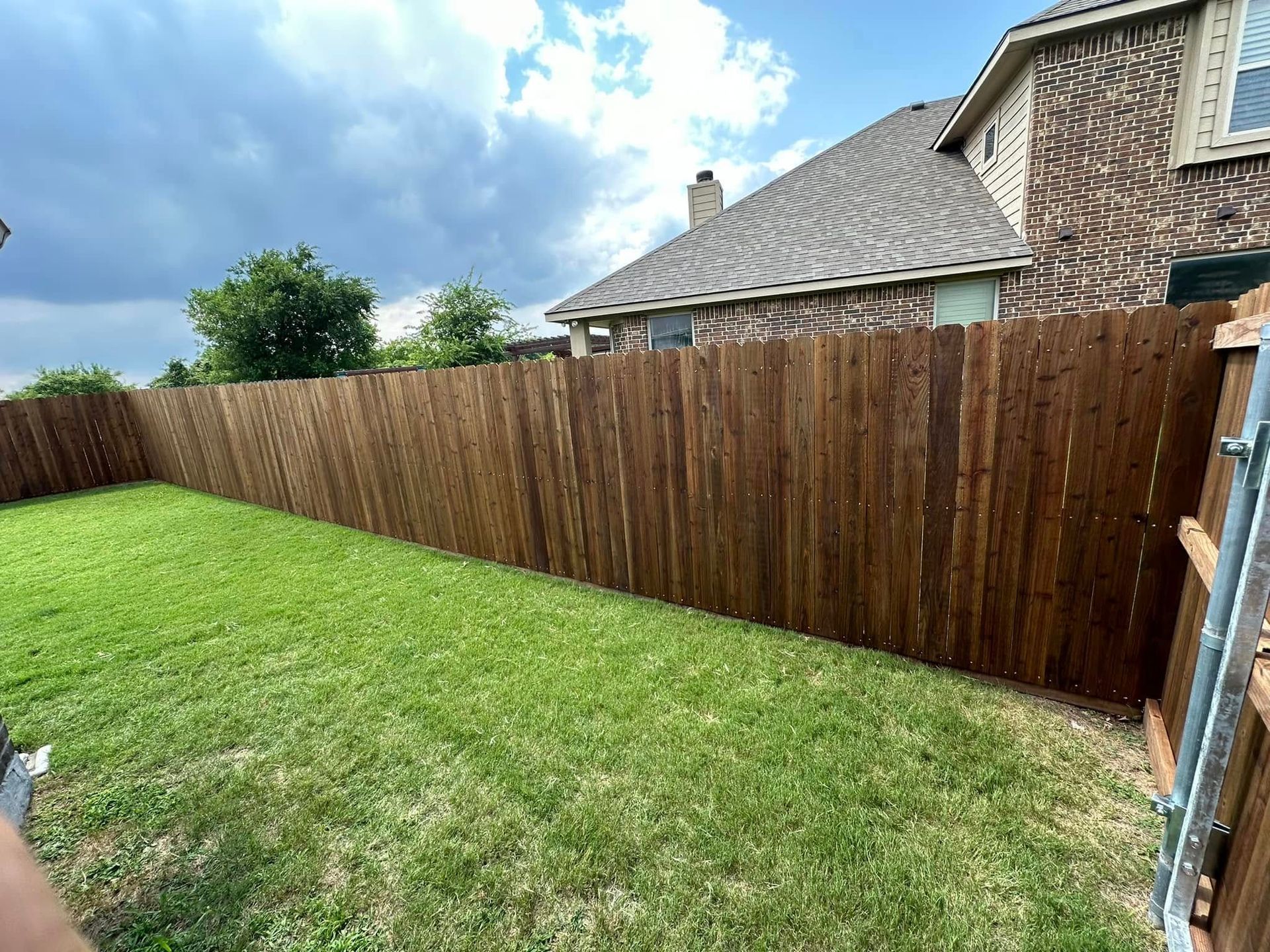 Backyard with green lawn, wooden privacy fence, and stone house under a cloudy sky