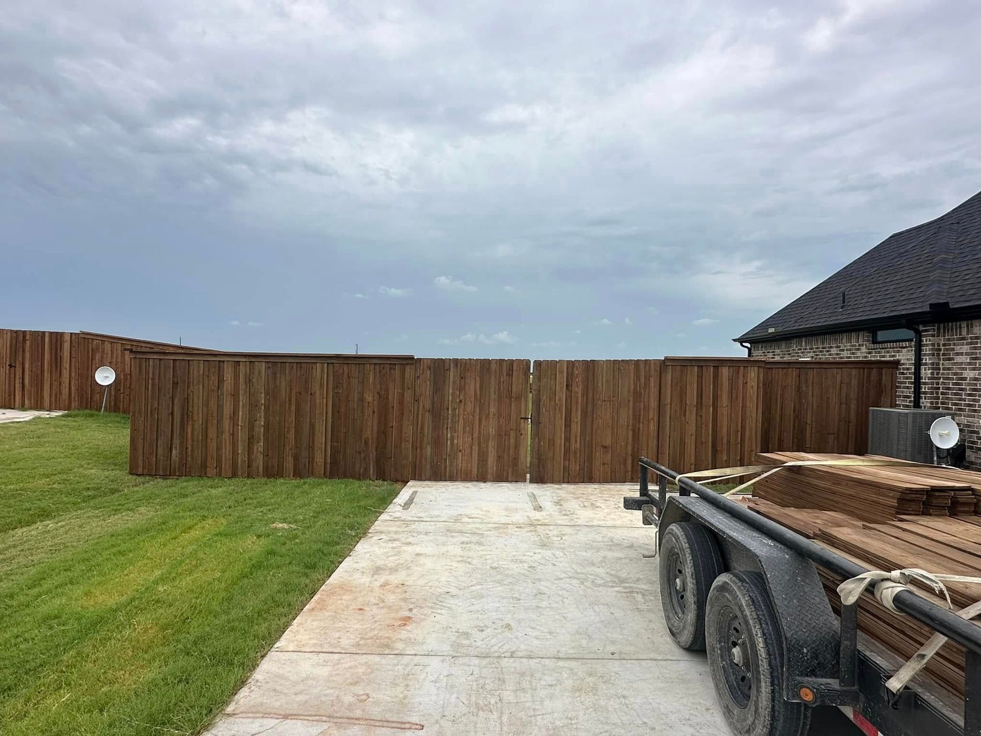 Wooden privacy fence behind a grassy yard, with a trailer and house on the right under a cloudy sky.
