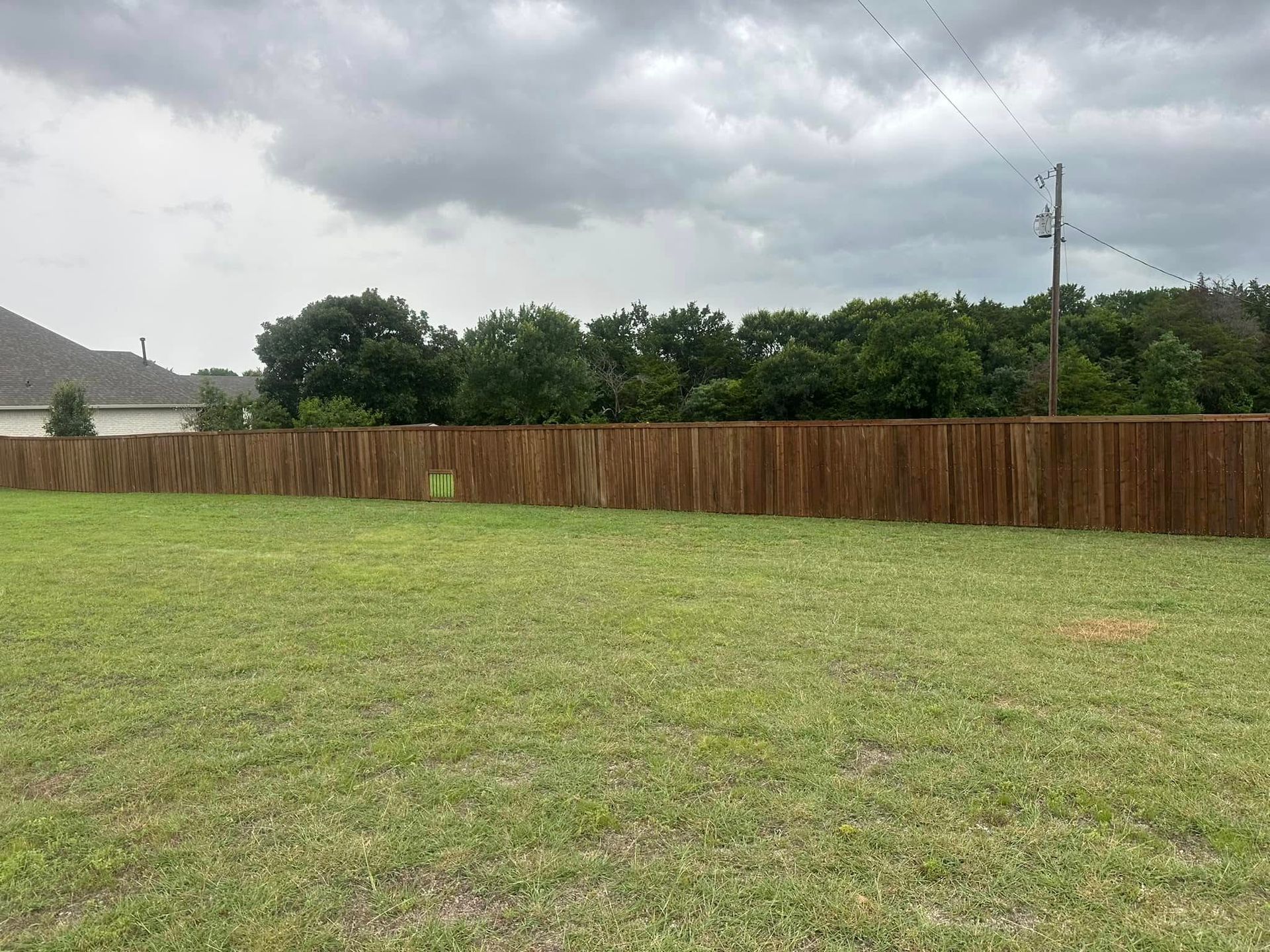 Grassy backyard with a wooden fence, trees, and cloudy sky