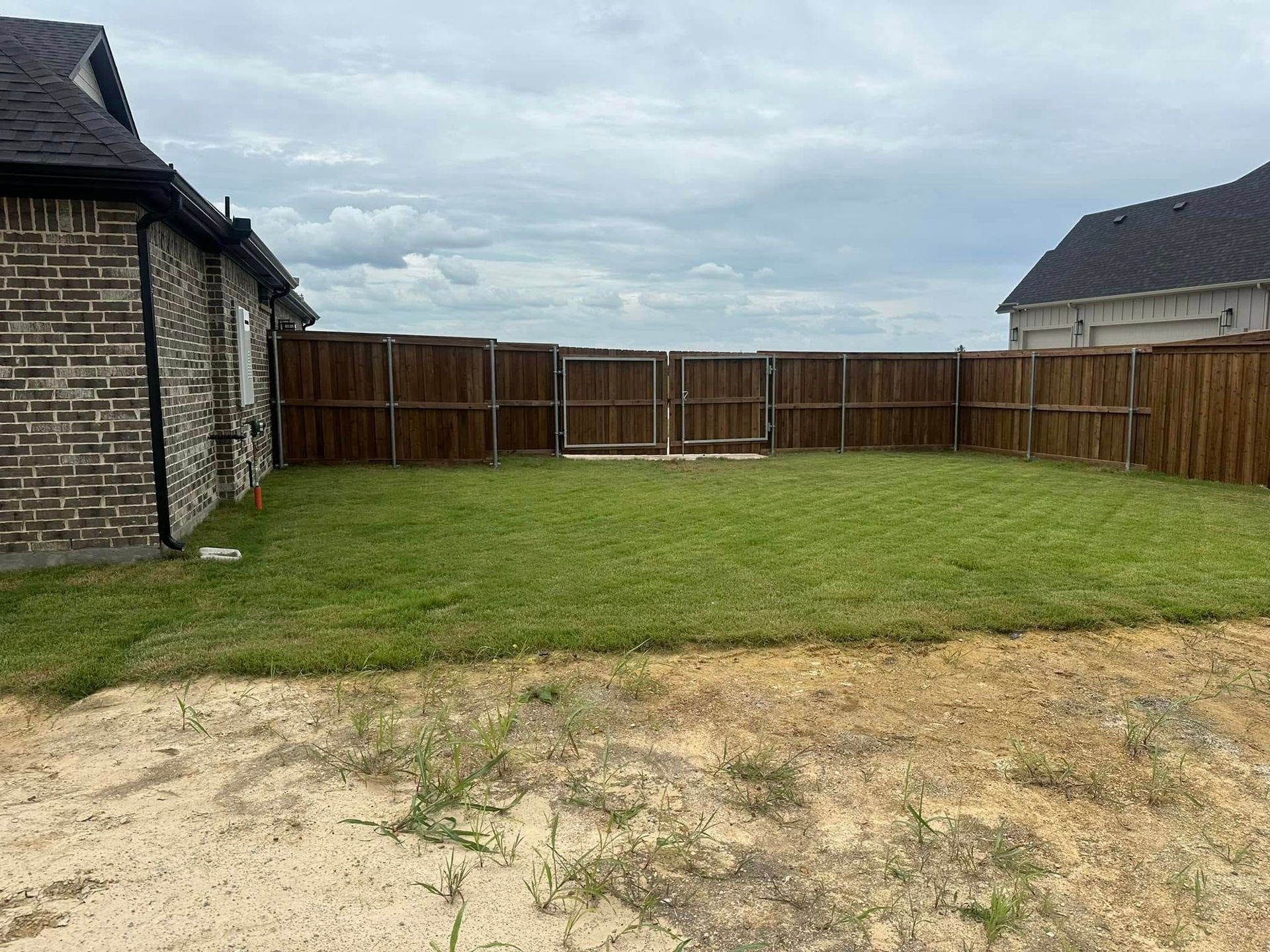 Backyard with grass, wooden fence, and brick houses under a cloudy sky