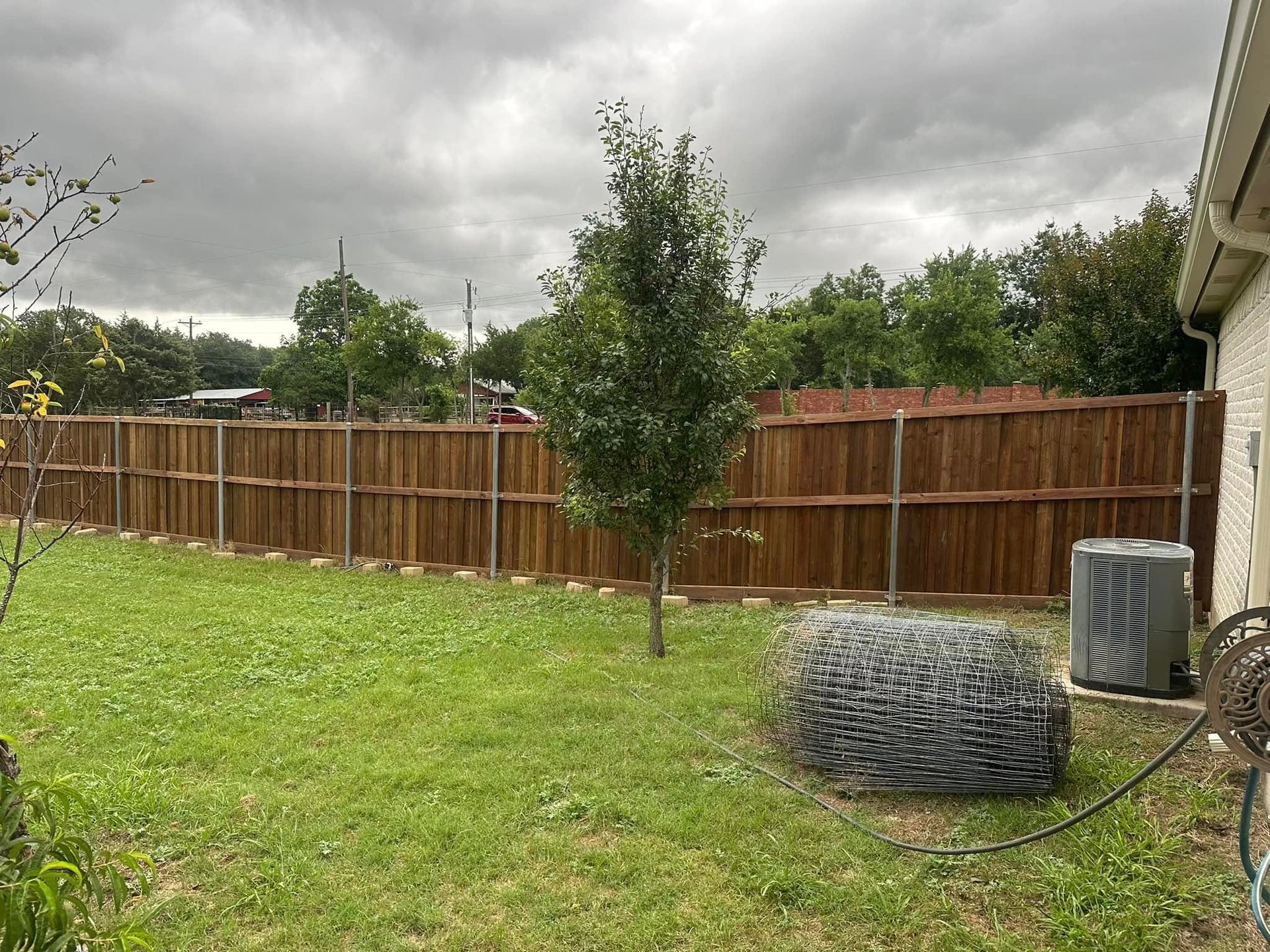 Backyard with green lawn, wooden fence, cloudy sky, small tree, and outdoor AC unit