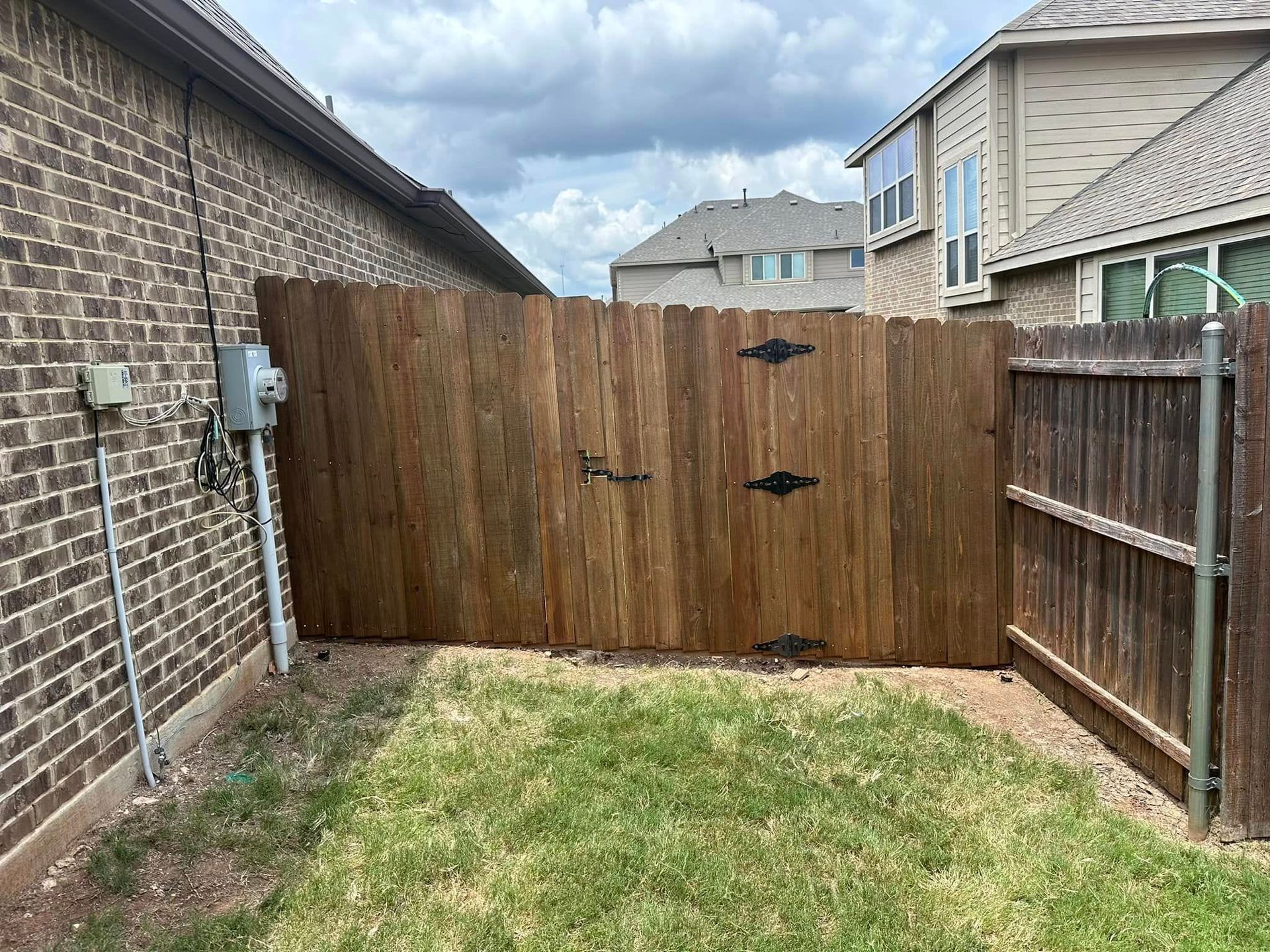 Small fenced backyard with a wooden gate, brick wall, and patchy grass under a cloudy sky.