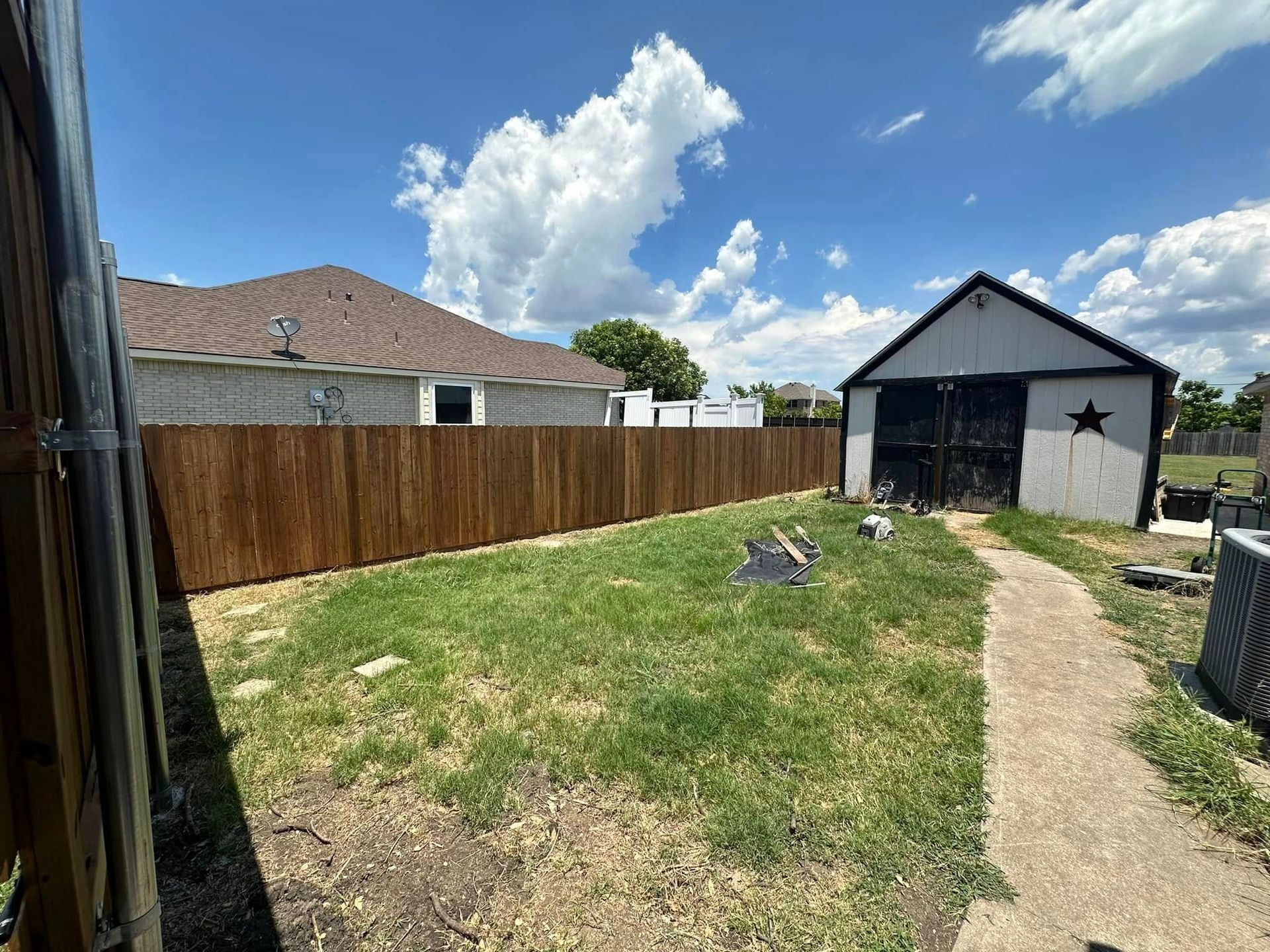 Backyard with brown fence, gravel path, grass, and a small shed under a blue sky.