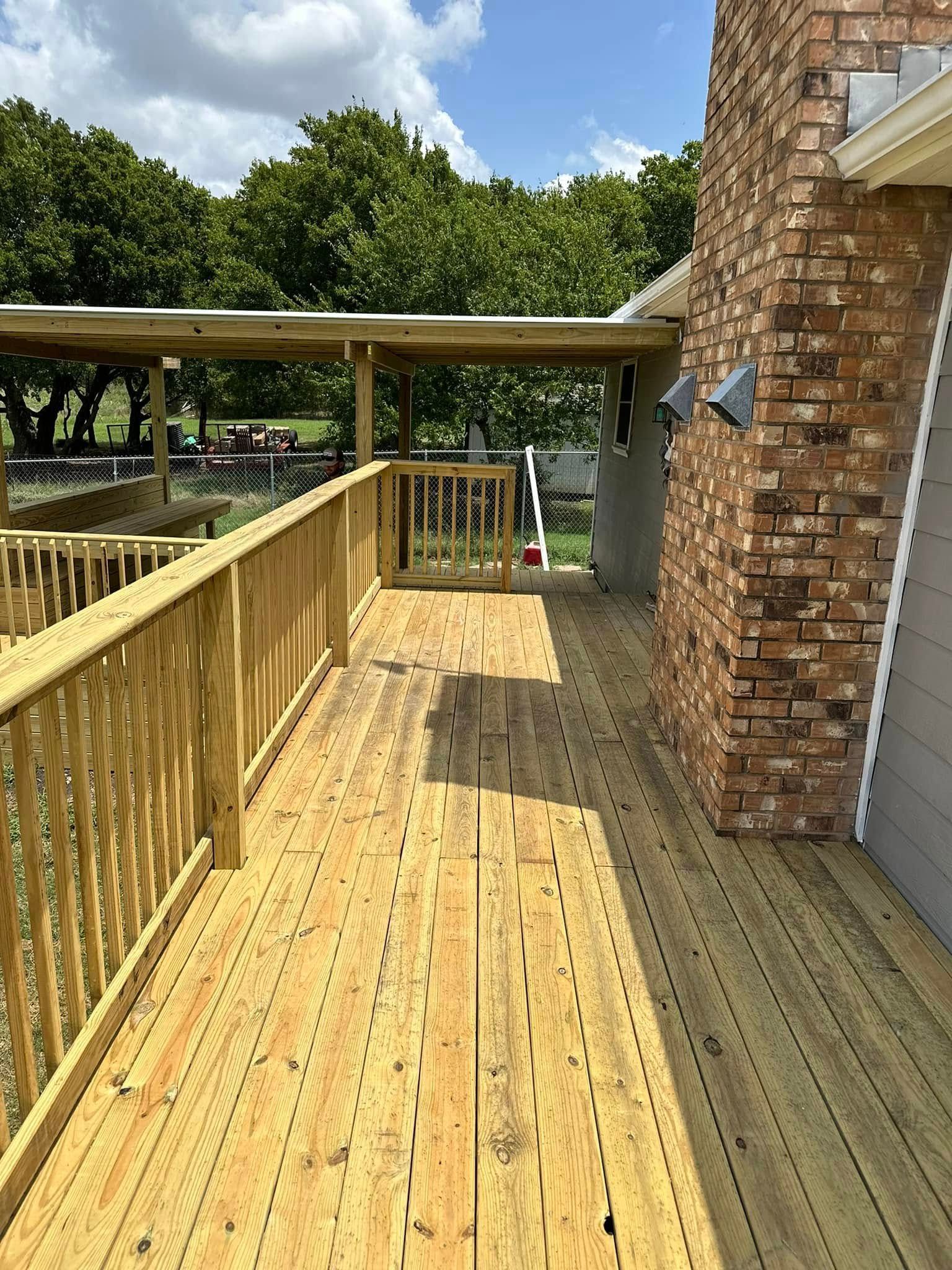 Wooden deck with yellow railing beside a brick house, leading to a covered patio and trees beyond.