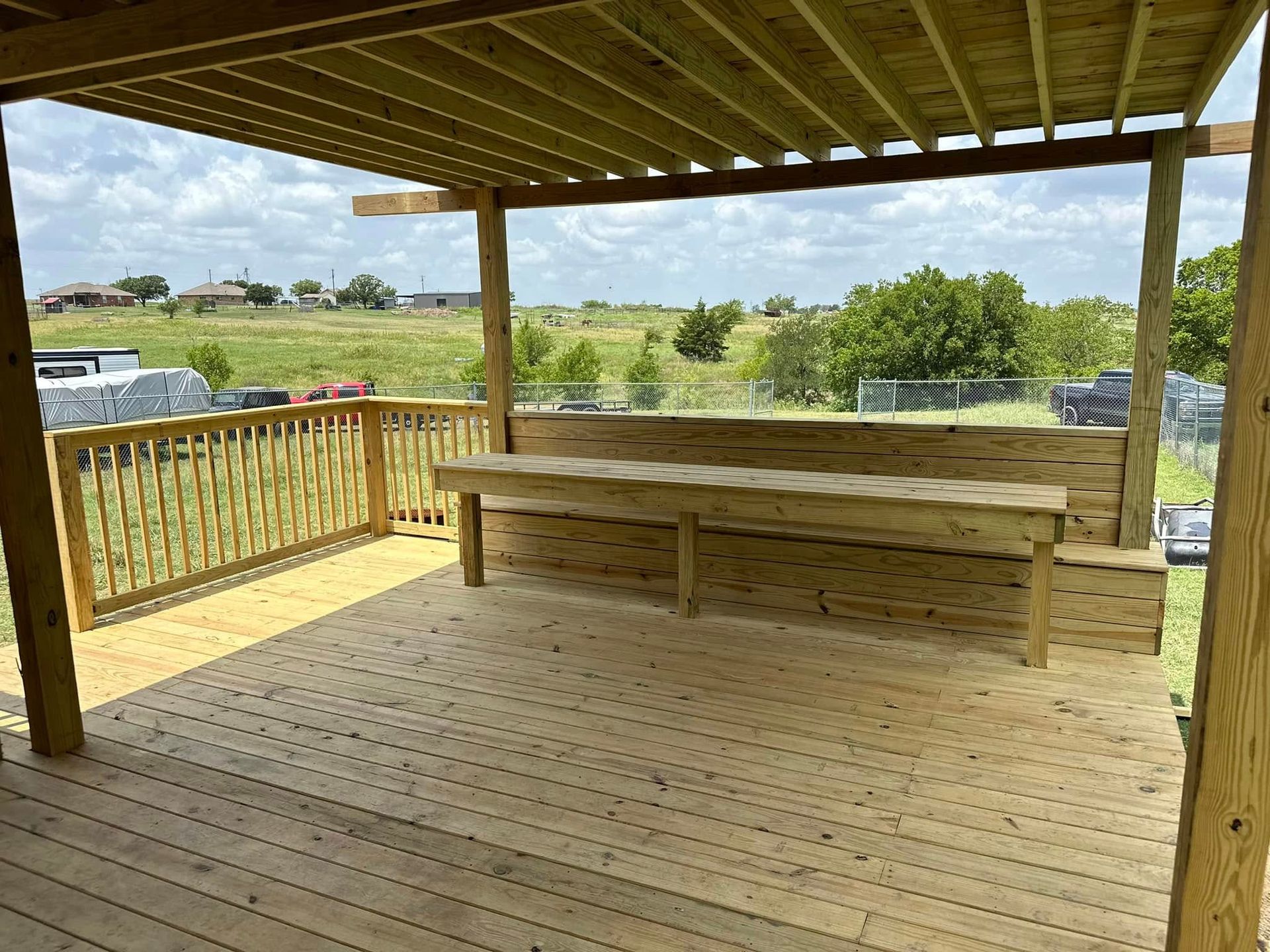 Covered wooden deck with railing, built-in bench, and view of open fields under a cloudy sky