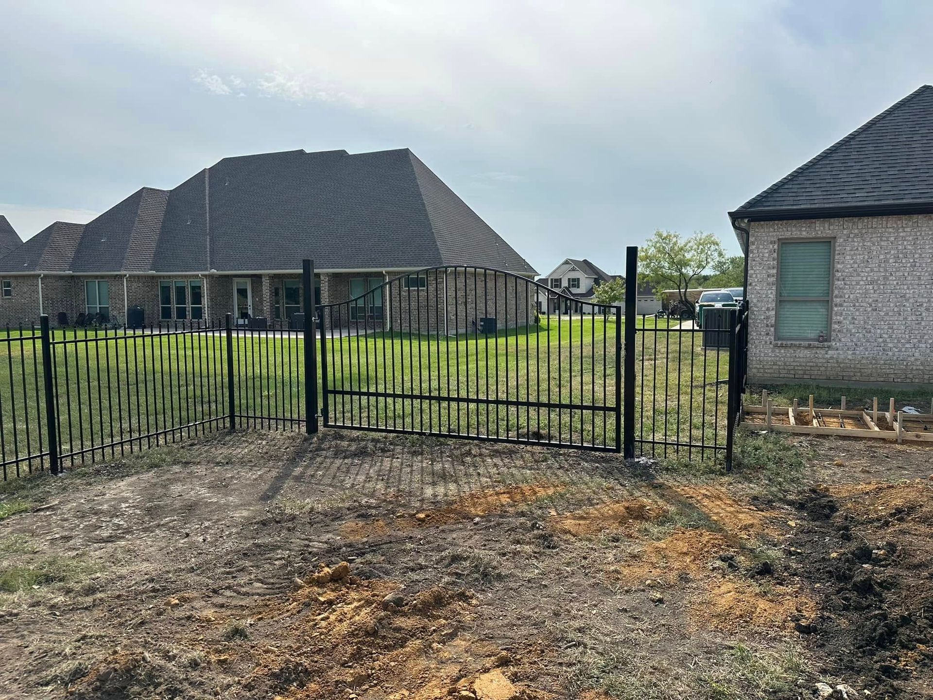 Black metal gate in a dirt lot, with houses and a fence behind it under a bright sky.