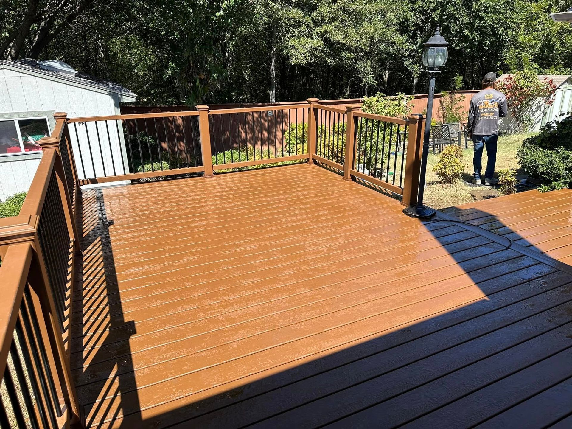 Wooden deck with railing in a sunny backyard, with one person standing near the garden edge.