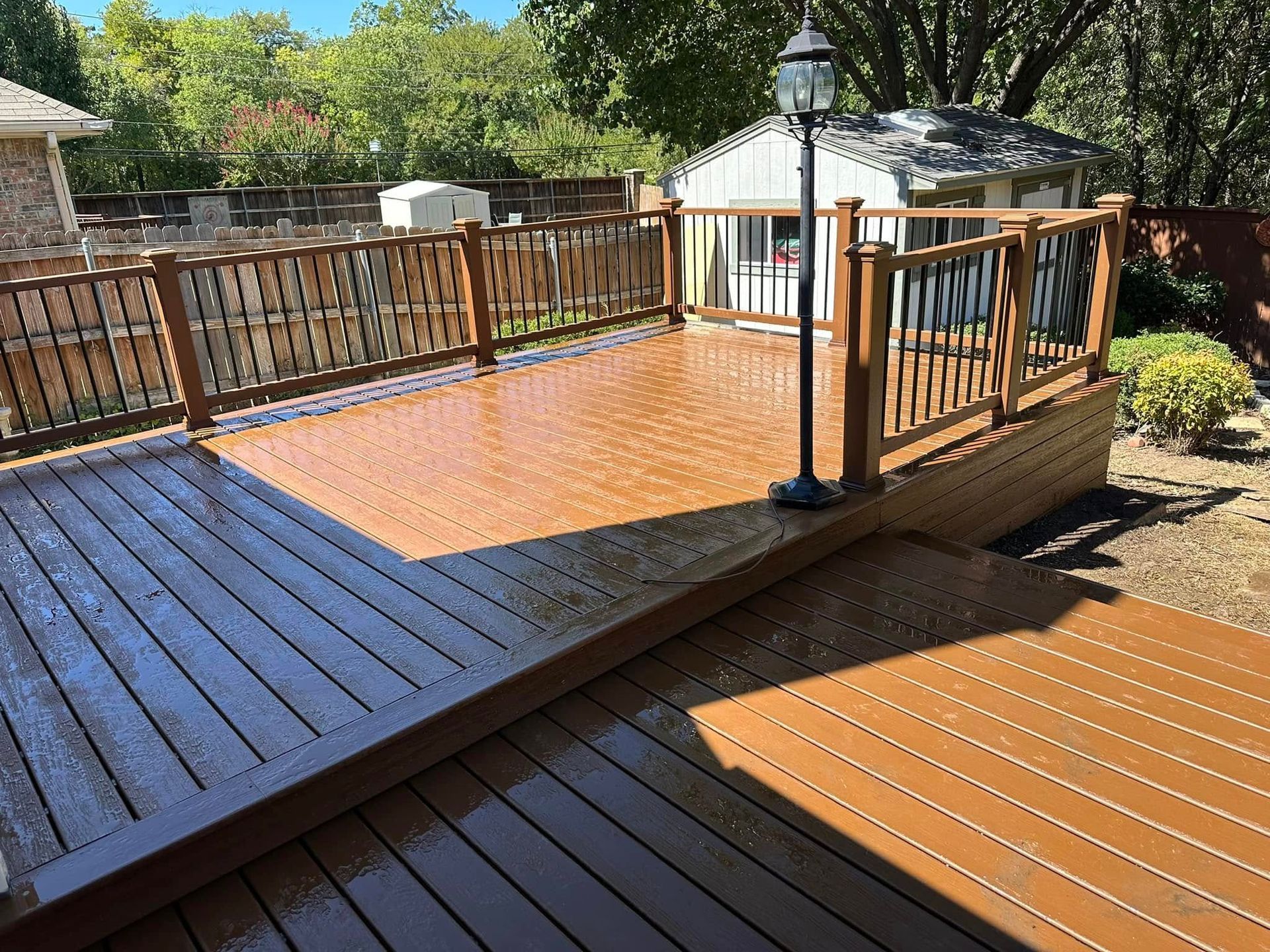 Wooden backyard deck with orange stain, railing, steps, and a lamppost in a sunny yard