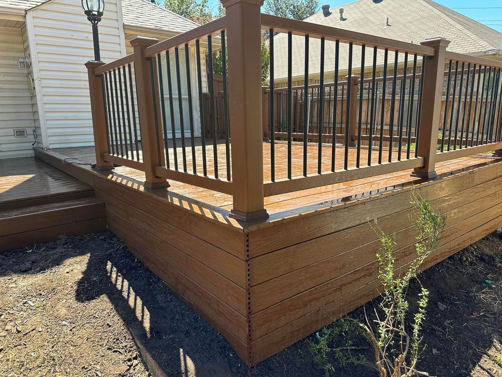 Wooden deck with brown railings and steps beside a house, surrounded by gravel and a small plant.
