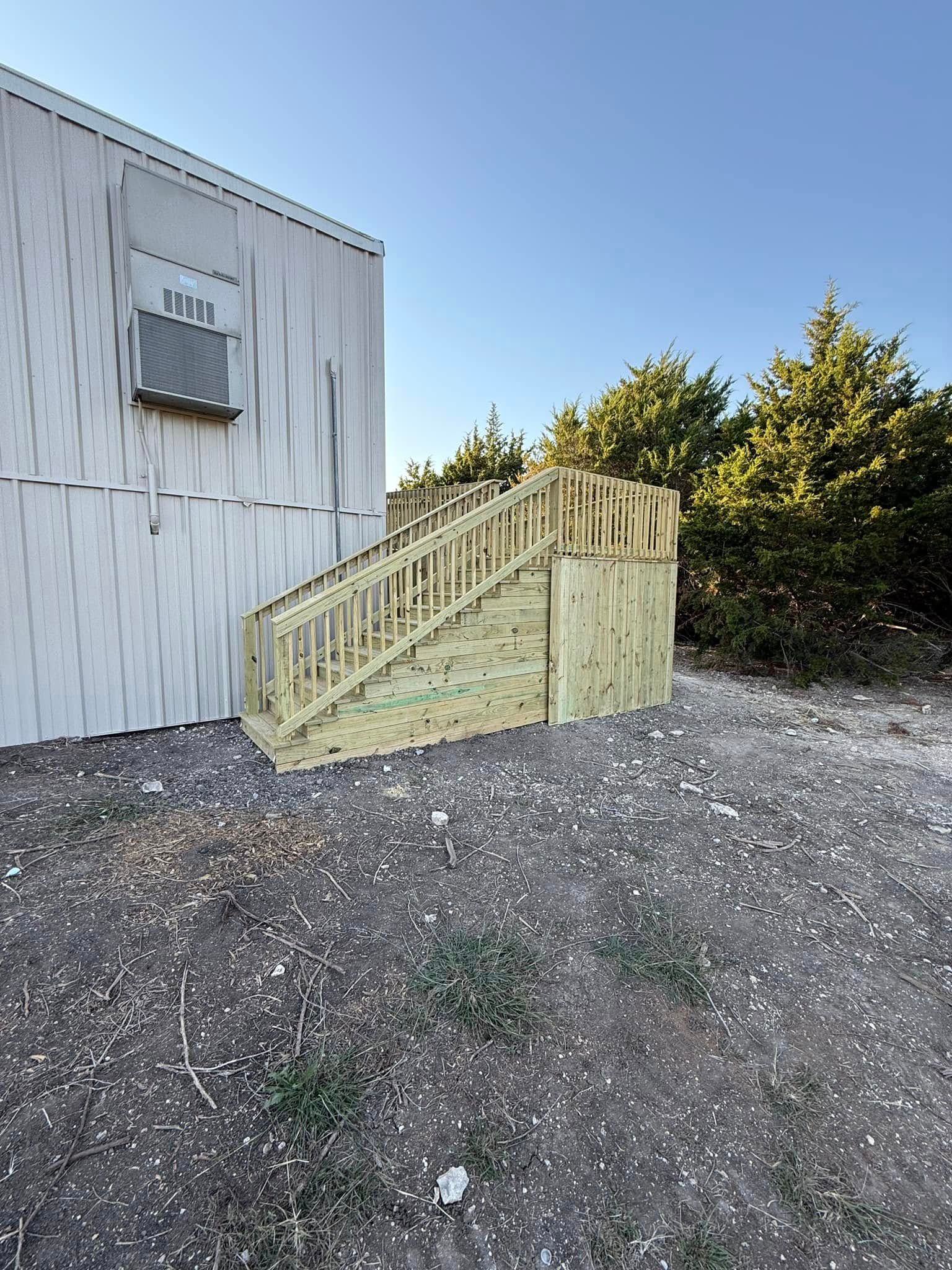 Wooden stairs beside a white building, with gravel ground and trees in the background.