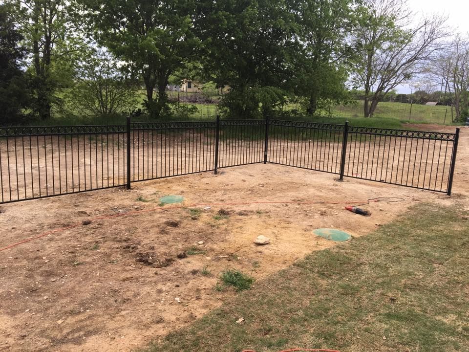 Dirt patch inside black metal fence in a grassy yard with trees in the background