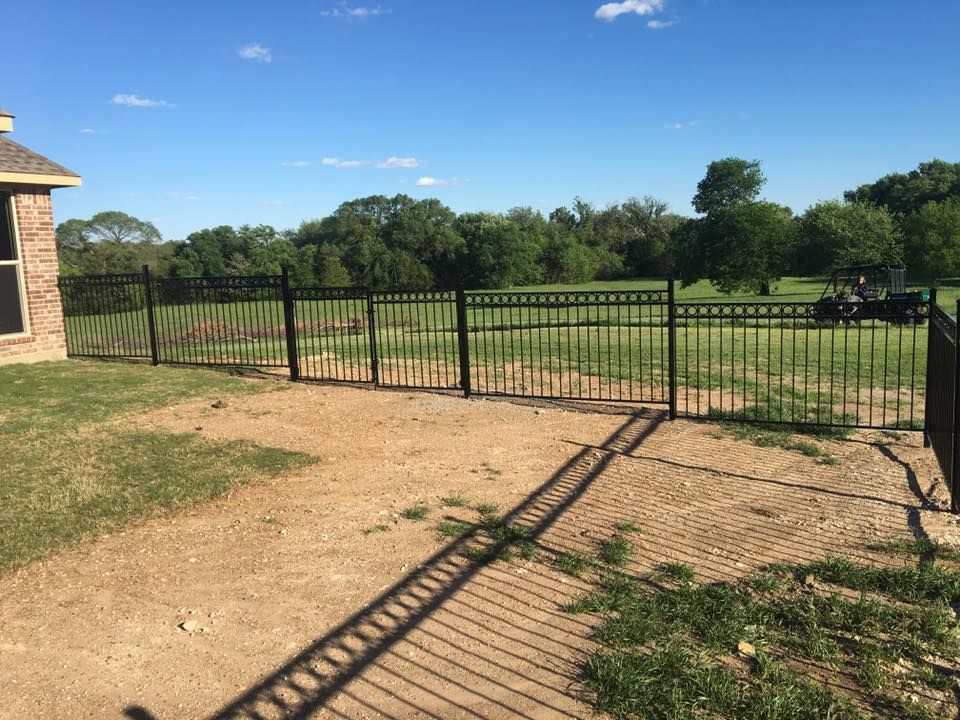 Black metal gate beside a grassy field, with a dirt path and trees under a clear blue sky