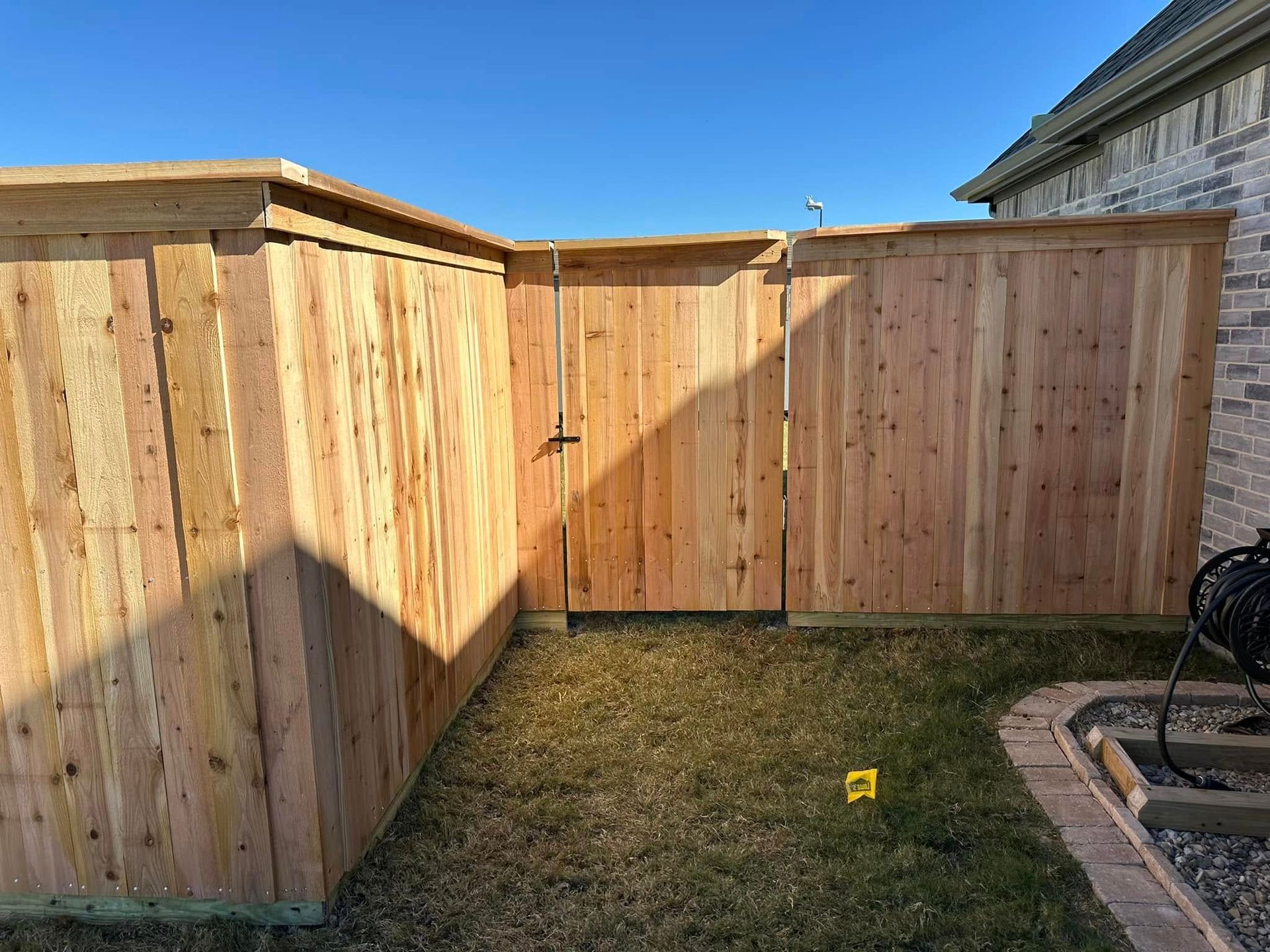Wooden backyard fence and shed-like enclosure beside a brick wall, with grass and a sunny sky.