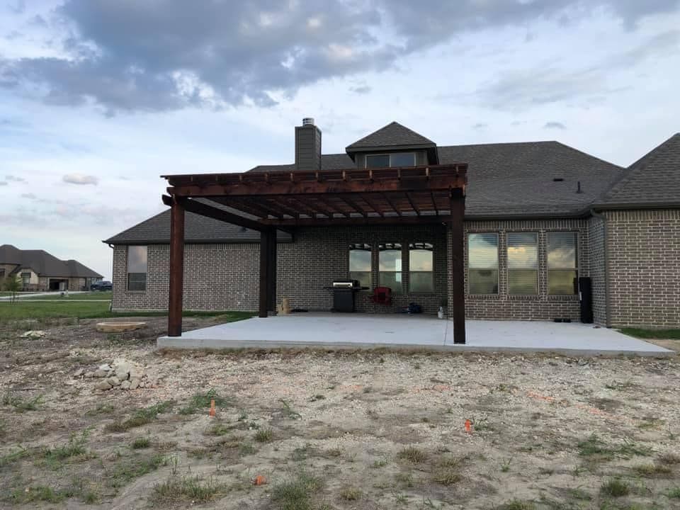 Backyard patio with a covered pergola beside a brick house and cloudy sky.