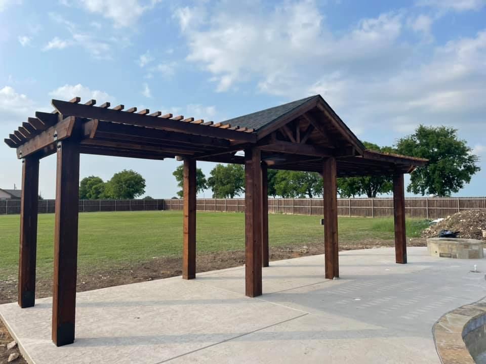 Dark wooden pergola on a concrete patio beside a grassy field under a cloudy sky