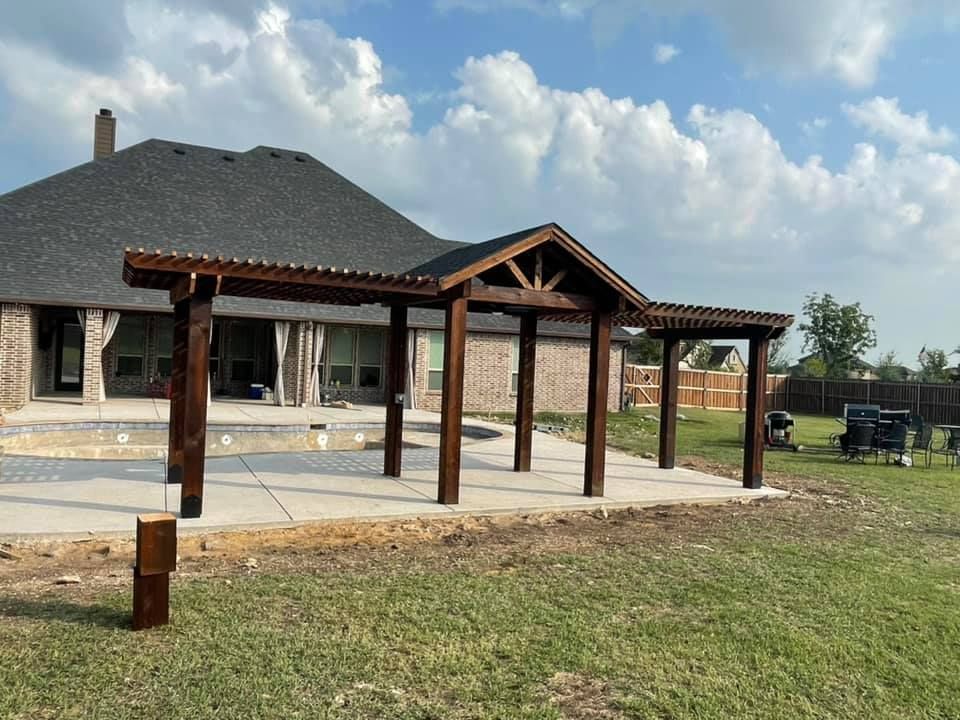 Covered patio with wooden pergola posts beside a house, on a concrete slab in a grassy yard under cloudy sky