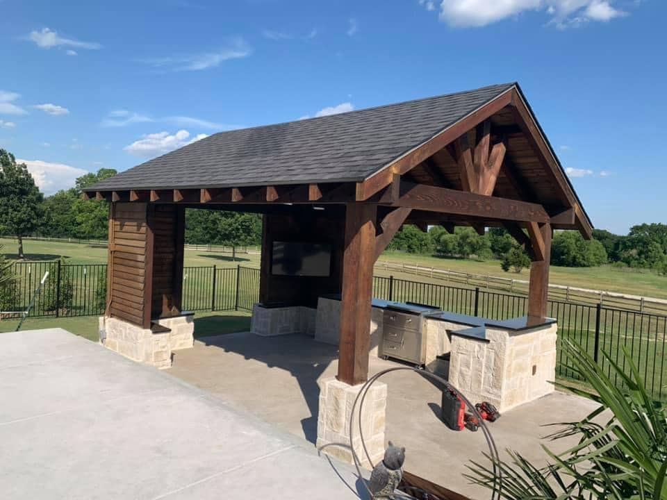 Wooden park pavilion with stone counters and red grill on a concrete pad, surrounded by grassy fields under blue sky