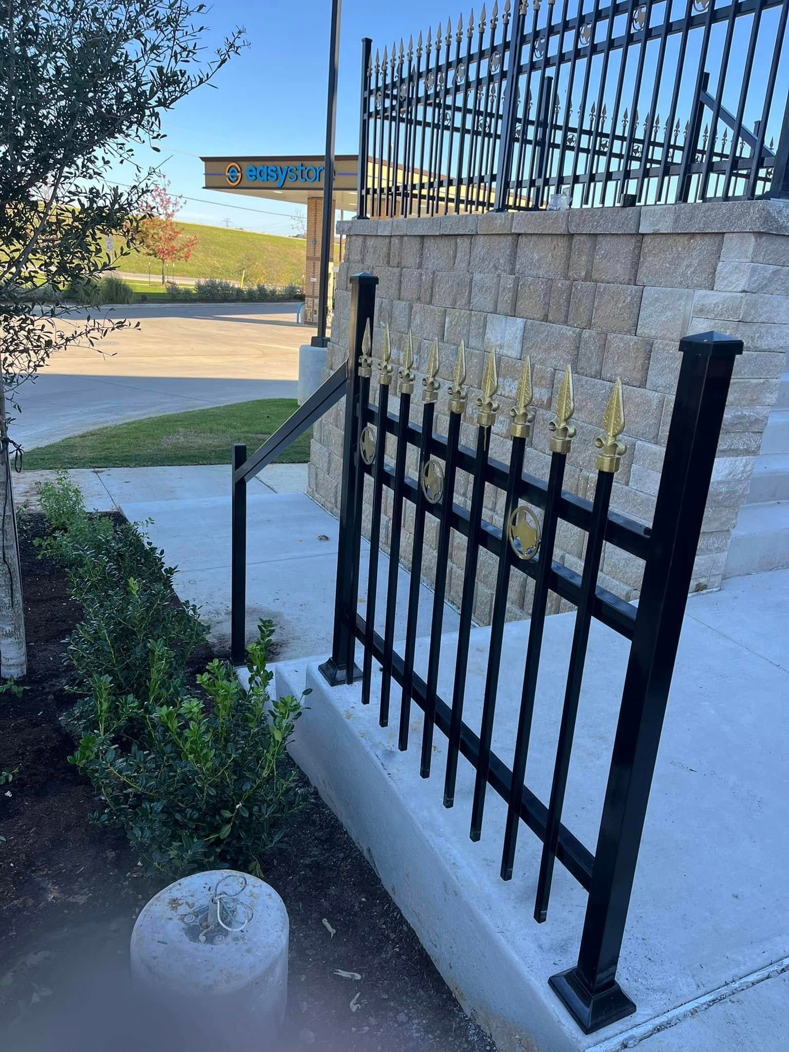 Black metal fence beside a white stucco wall overlooking a sunny roadside entrance