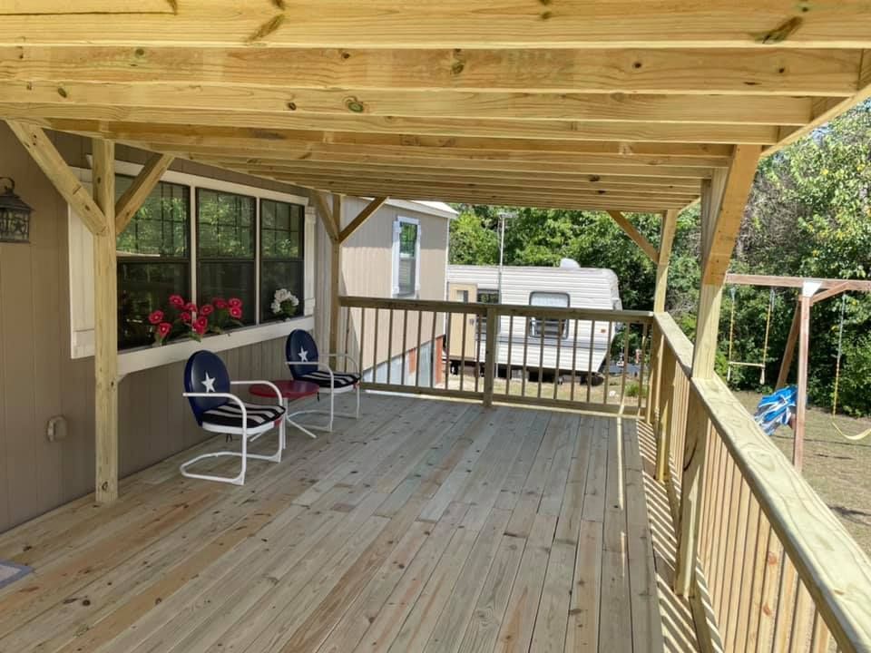 Covered wooden deck with railing, two chairs, and a window box of red flowers beside a house.